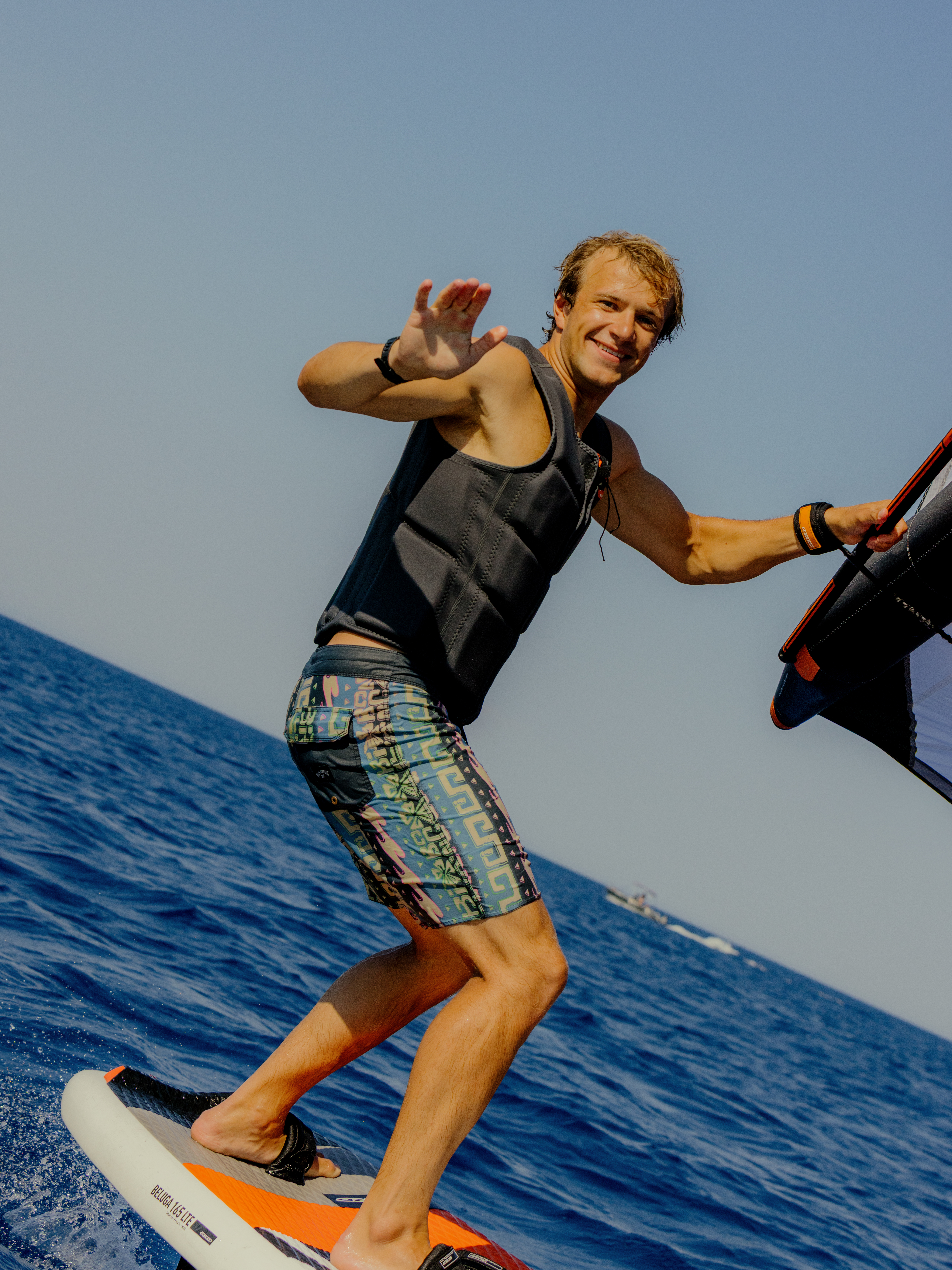 A young man wakeboarding on calm blue water, wearing a black life vest and colorful patterned board shorts. He's smiling at the camera while holding onto a tow rope with one hand and waving with the other. The sky is clear and blue, and he appears to be mid-ride on an orange and white wakeboard.