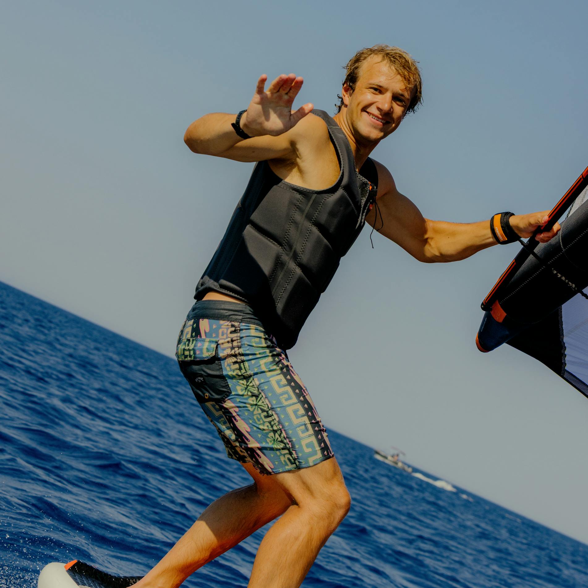 A young man wakeboarding on calm blue water, wearing a black life vest and colorful patterned board shorts. He's smiling at the camera while holding onto a tow rope with one hand and waving with the other. The sky is clear and blue, and he appears to be mid-ride on an orange and white wakeboard.