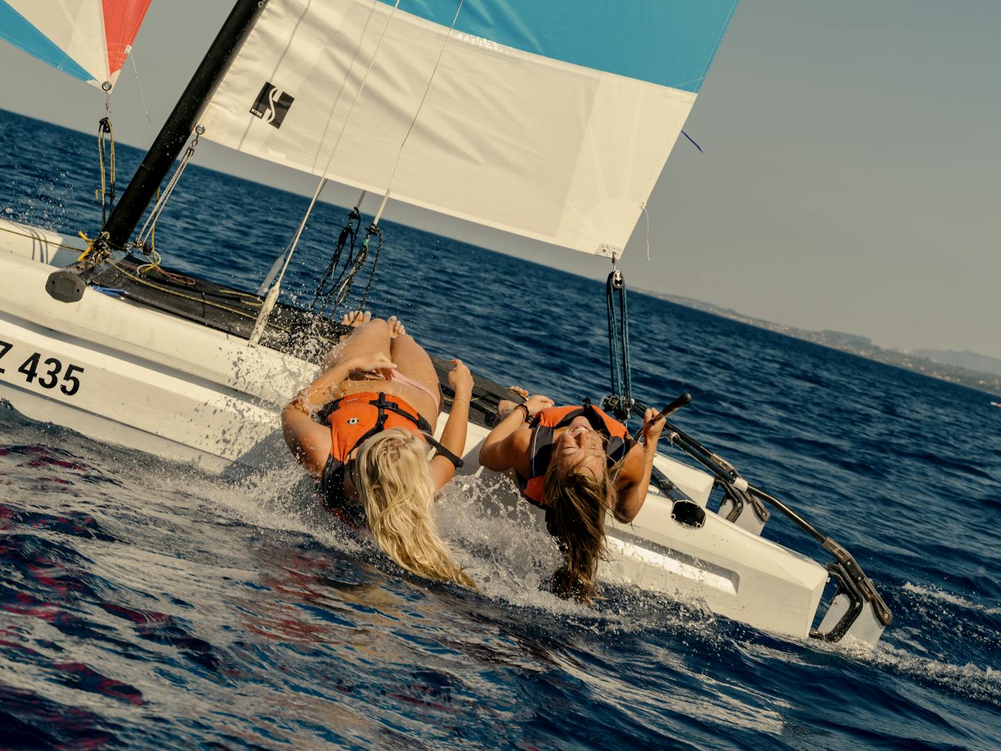 Two people sailing a small catamaran numbered 435, leaning back over the water to balance the boat as it moves at speed. The sailboat has colorful sails in red, cream, turquoise, and blue patterns with a black mast. Both sailors are wearing orange life vests and the boat is creating white spray as it cuts through the deep blue ocean water against a clear sky.