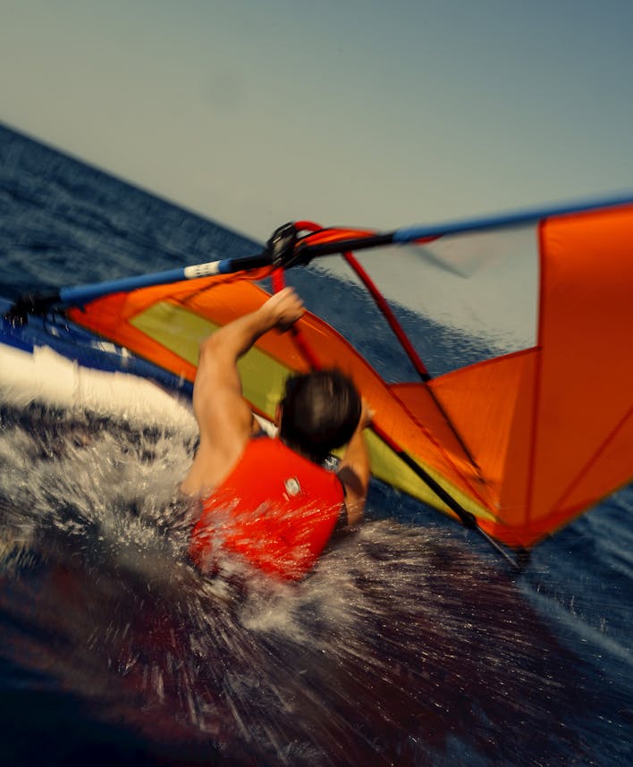 Action shot of a windsurfer navigating through choppy water, viewed from above. The rider wears red shorts and grips a colorful windsurf sail featuring orange, yellow, blue, and red panels. Water sprays dramatically around the board as it cuts through the dark blue ocean waves, creating white foam and motion blur that conveys speed and dynamic movement.