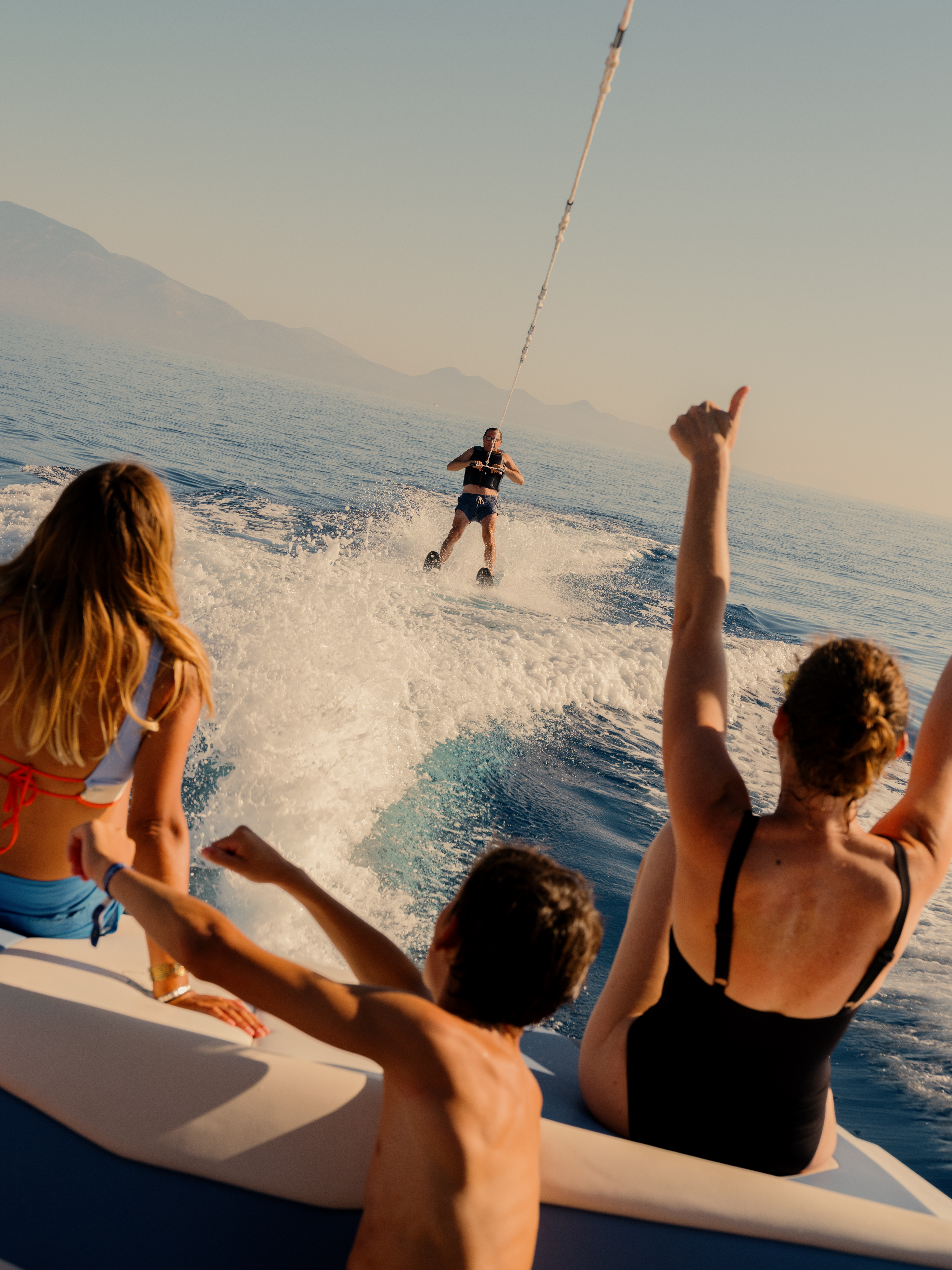 A group of people on a boat watching and cheering as a wakeboarder rides behind them, creating a white spray of water. Three spectators in the foreground have their arms raised in excitement while viewing from the boat's stern. Mountains are visible in the hazy background across the calm blue water during golden hour lighting.