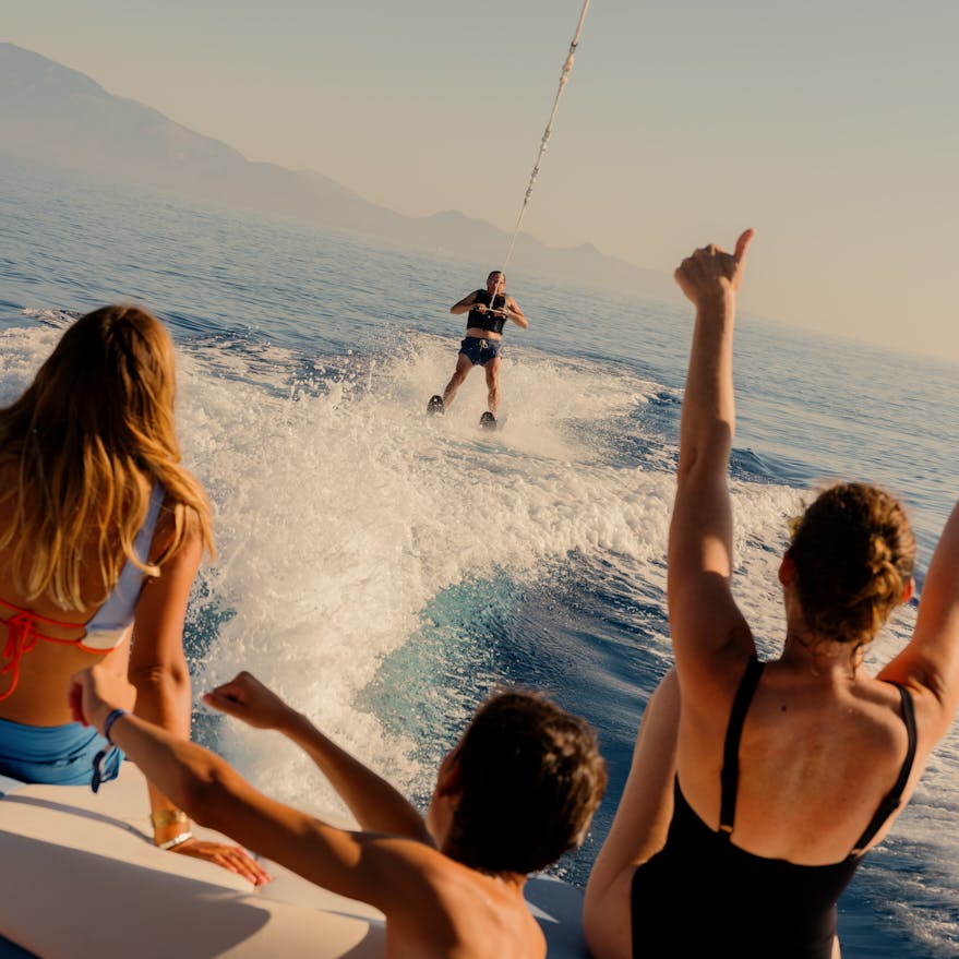 A group of people on a boat watching and cheering as a wakeboarder rides behind them, creating a white spray of water. Three spectators in the foreground have their arms raised in excitement while viewing from the boat's stern. Mountains are visible in the hazy background across the calm blue water during golden hour lighting.