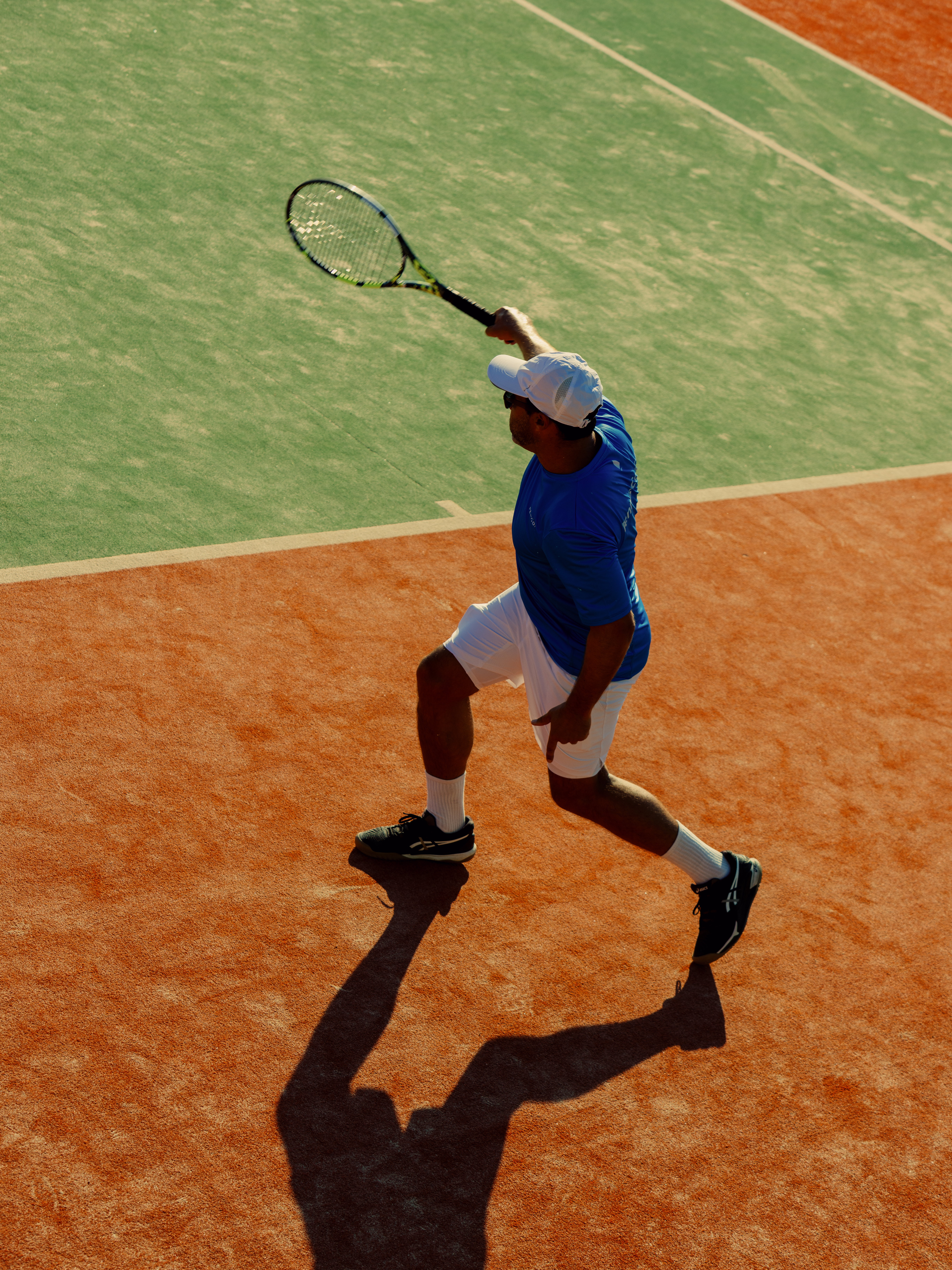 Overhead view of a tennis player mid-swing on a clay court, wearing a blue shirt, white shorts, white cap, and black shoes. The player holds a tennis racket raised above their head, casting a distinct shadow on the orange-red clay surface. The adjacent grass court is visible in the background, creating a contrast between the two playing surfaces.