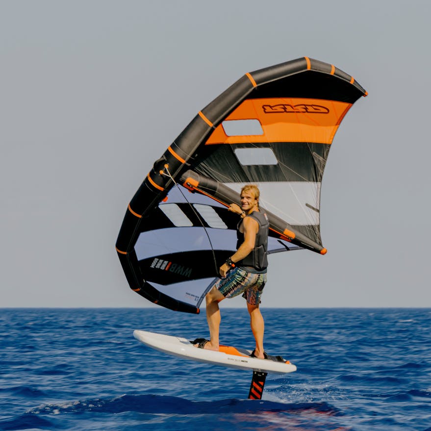 A wing foiler rides above the water on a hydrofoil board, holding a large wing sail with orange, gray, and black stripes. The rider wears a black vest and patterned shorts while gliding over calm, deep blue ocean water against a clear sky.