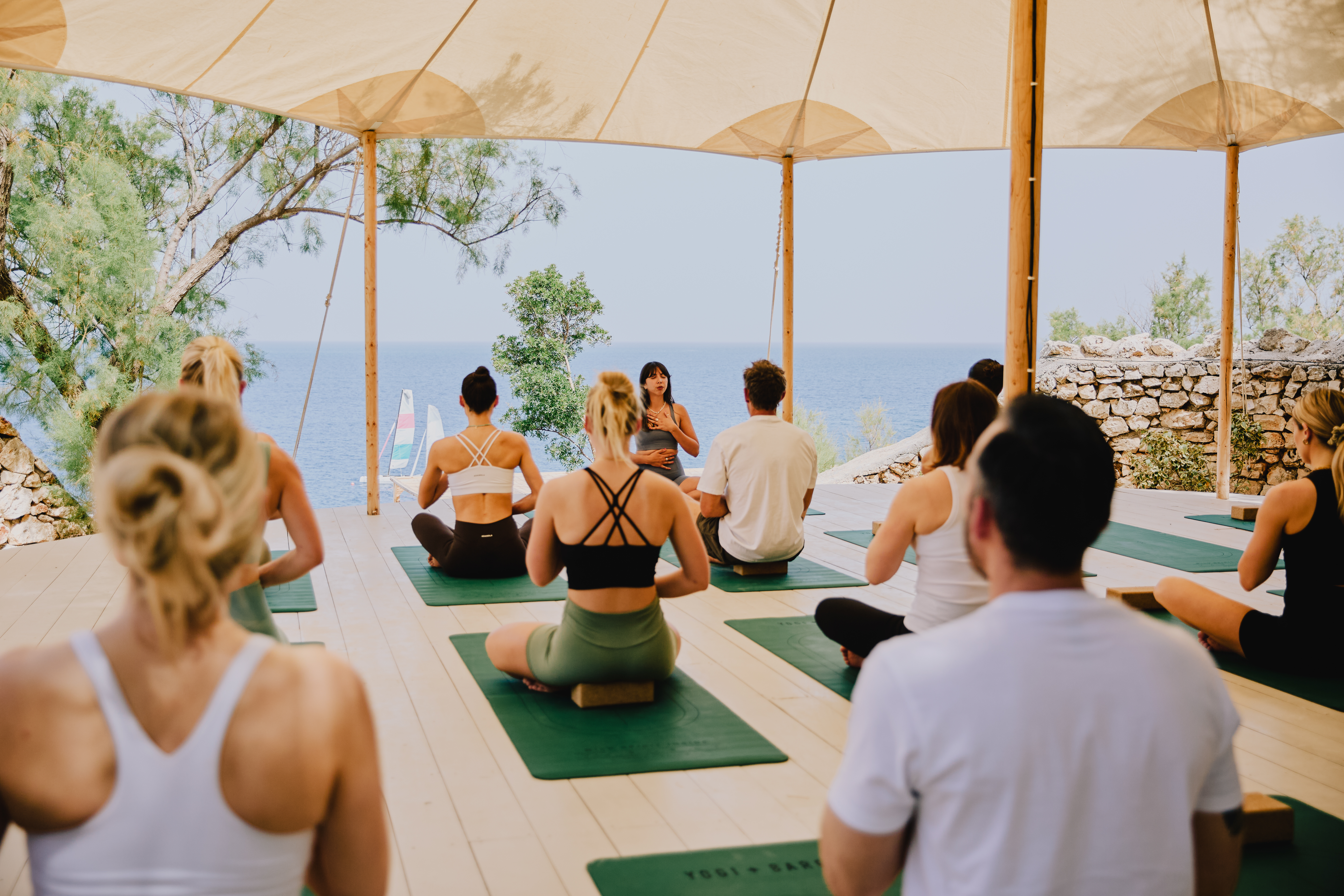 A yoga instructor leads an outdoor class under a large beige canopy with multiple participants seated on green yoga mats. The open-air pavilion overlooks the ocean with stone walls and natural vegetation surrounding the space. Participants wear athletic attire in white, black, and neutral tones while sitting in meditation poses facing the instructor and scenic coastal view.
