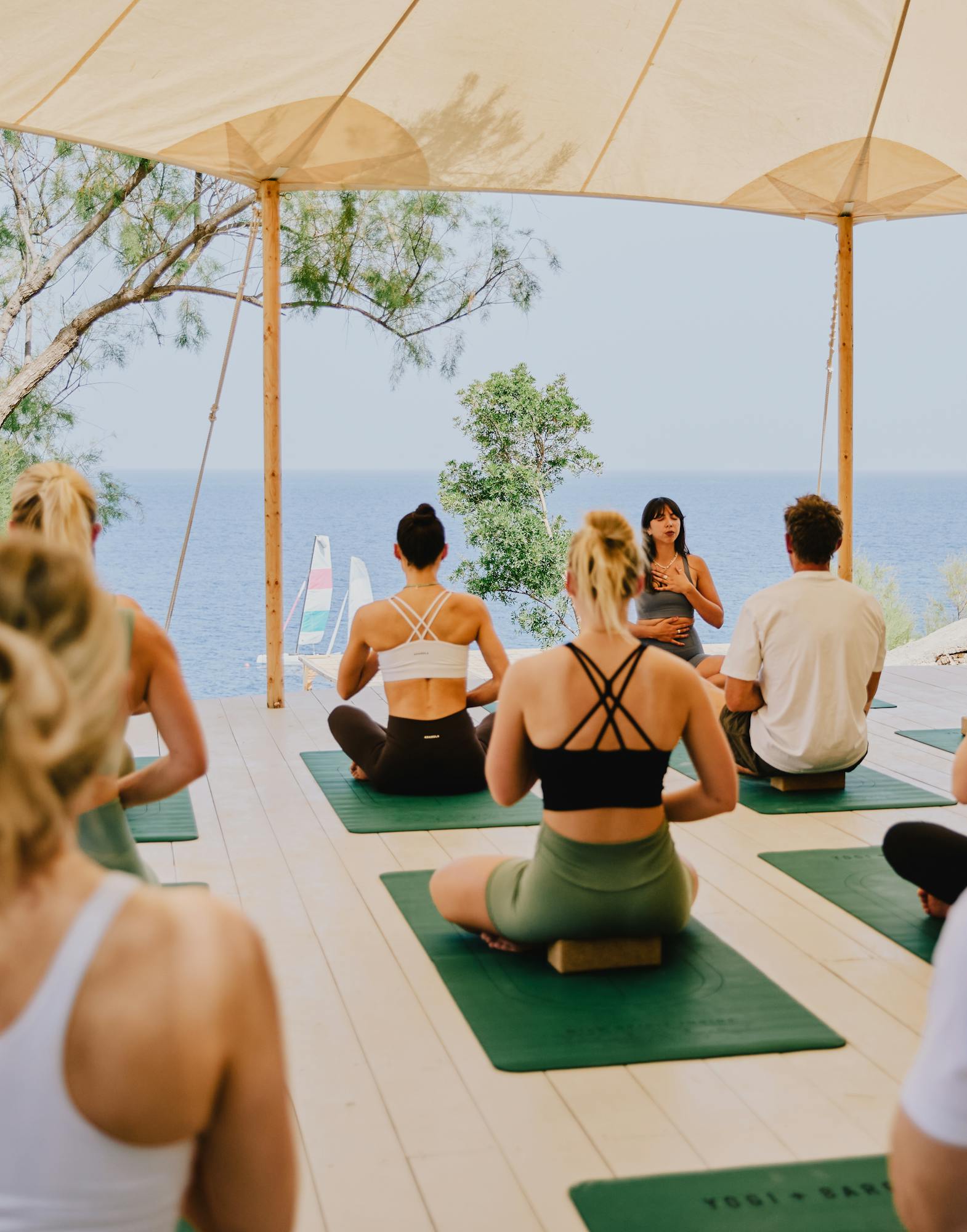 A yoga instructor leads an outdoor class under a large beige canopy with multiple participants seated on green yoga mats. The open-air pavilion overlooks the ocean with stone walls and natural vegetation surrounding the space. Participants wear athletic attire in white, black, and neutral tones while sitting in meditation poses facing the instructor and scenic coastal view.