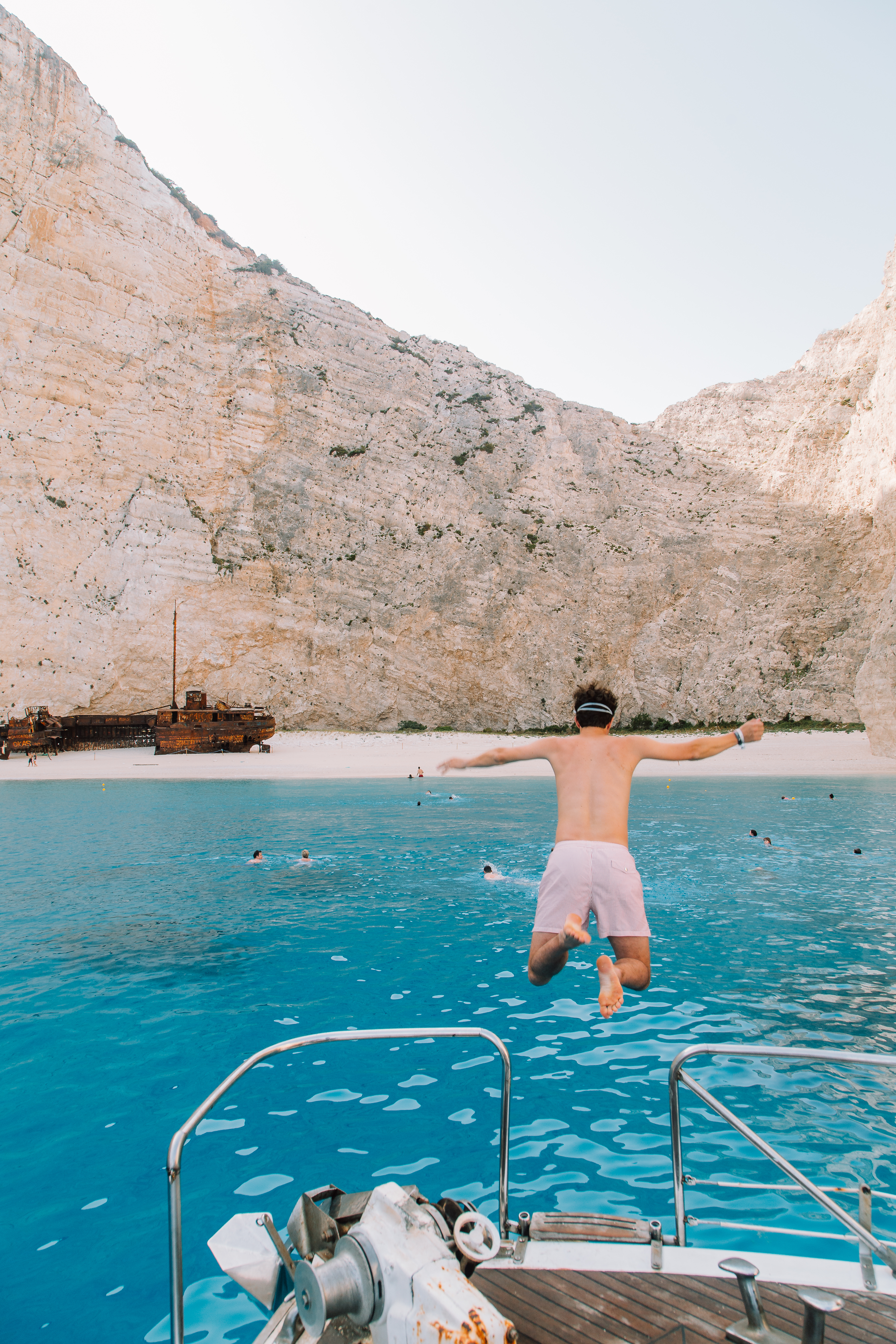 A shirtless man wearing striped shorts and a headband jumps off the back of a boat into brilliant turquoise water at Navagio Beach (Shipwreck Beach) in Zakynthos, Greece. The dramatic scene shows towering limestone cliffs framing the secluded cove, with a famous rusted shipwreck visible on the white sand beach. Several other swimmers can be seen in the crystal-clear water