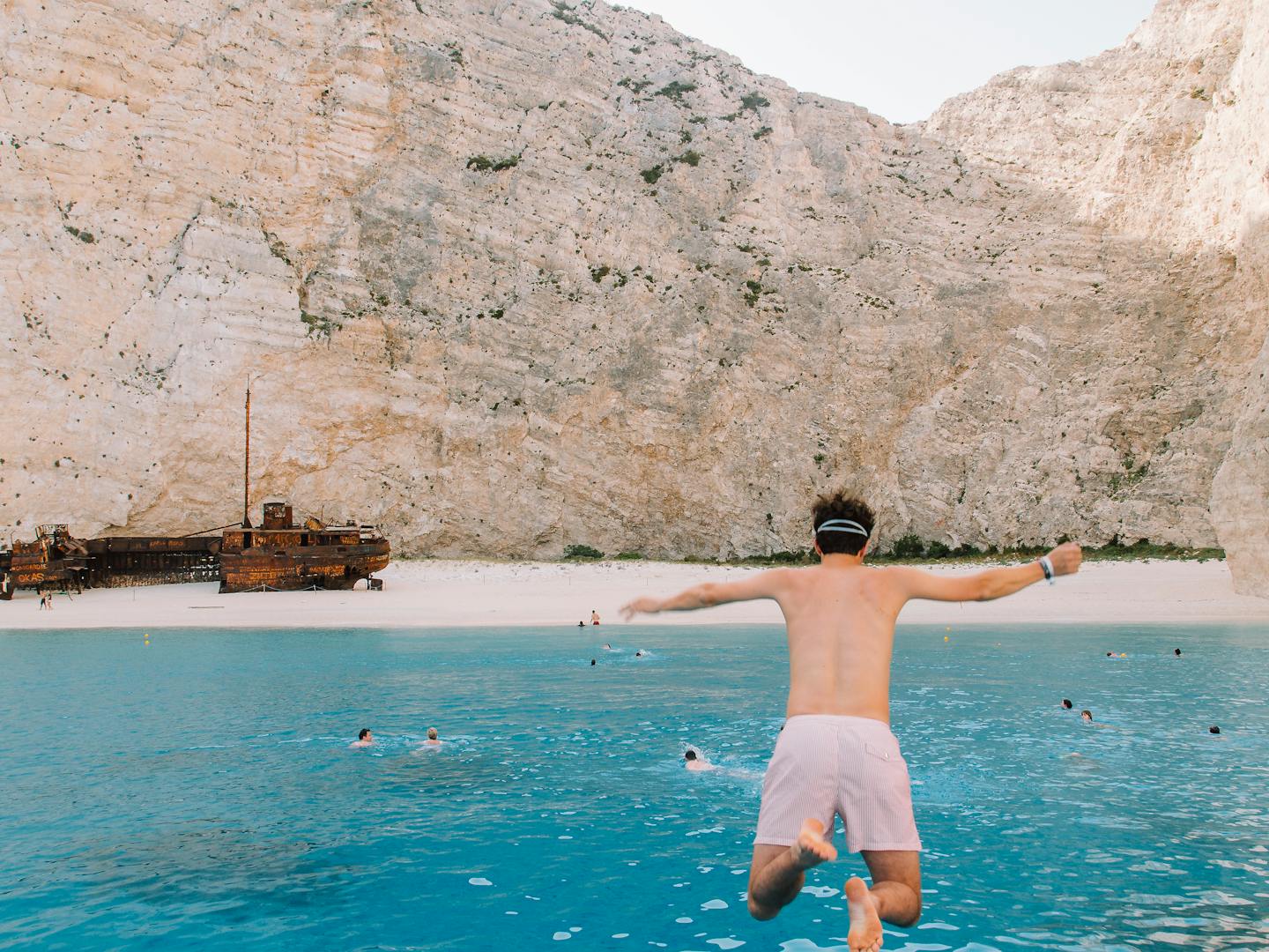 A shirtless man wearing striped shorts and a headband jumps off the back of a boat into brilliant turquoise water at Navagio Beach (Shipwreck Beach) in Zakynthos, Greece. The dramatic scene shows towering limestone cliffs framing the secluded cove, with a famous rusted shipwreck visible on the white sand beach. Several other swimmers can be seen in the crystal-clear water