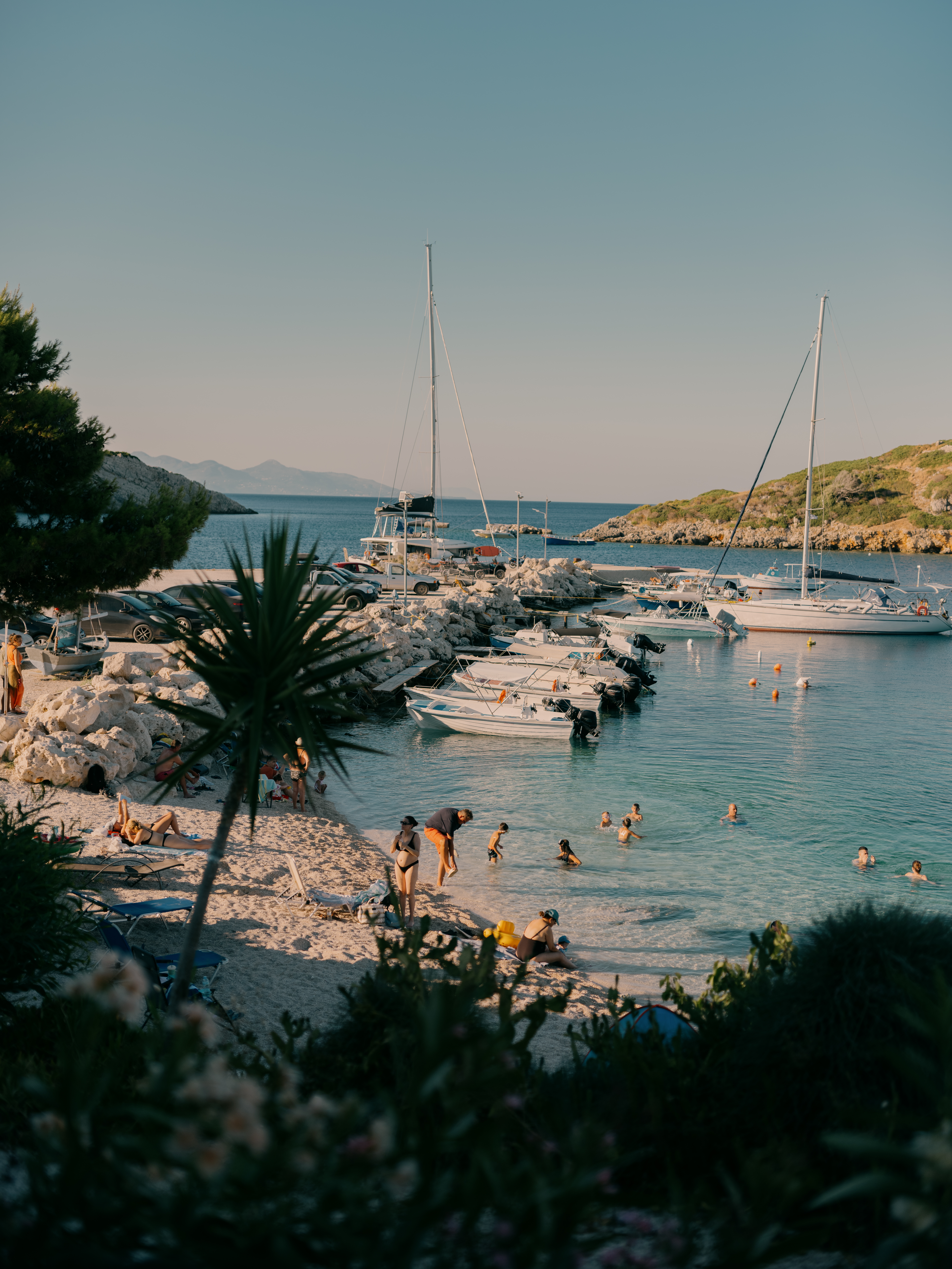 A scenic Mediterranean coastal cove during golden hour, featuring a small marina with sailboats and smaller vessels docked along a rocky breakwater. Beachgoers wade in the calm turquoise water while others relax on the sandy beach. Palm trees and lush vegetation frame the scene, with mountains visible across the tranquil sea in the distance under a clear sky.