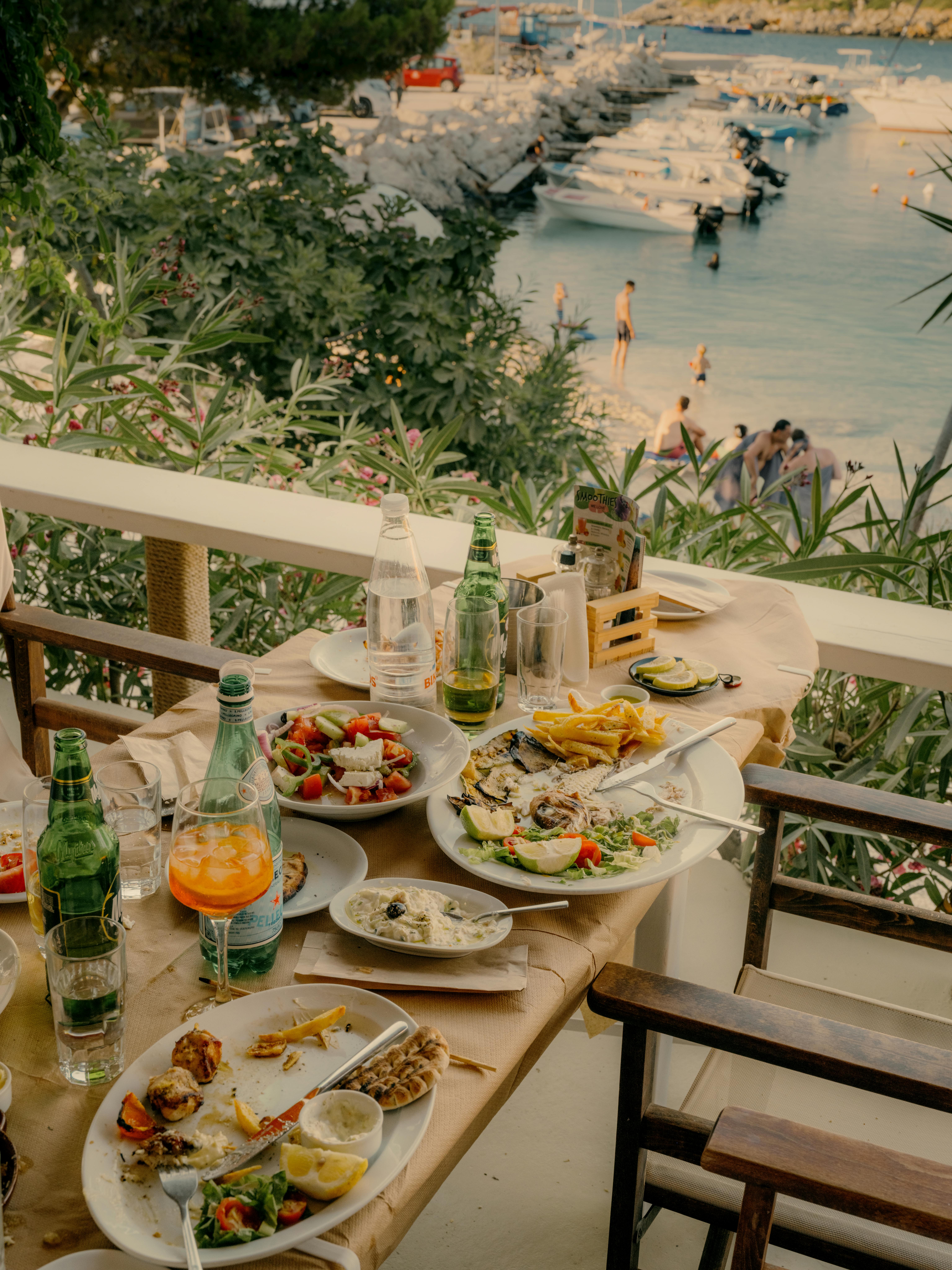 An outdoor waterfront dining table set with Mediterranean cuisine including Greek salad, grilled fish with lemon, french fries, and seafood appetizers. Green glass bottles and colorful drinks accompany the meal. Beyond the white railing with lush greenery, a marina with white boats is visible, and beachgoers can be seen in the background during golden hour lighting.