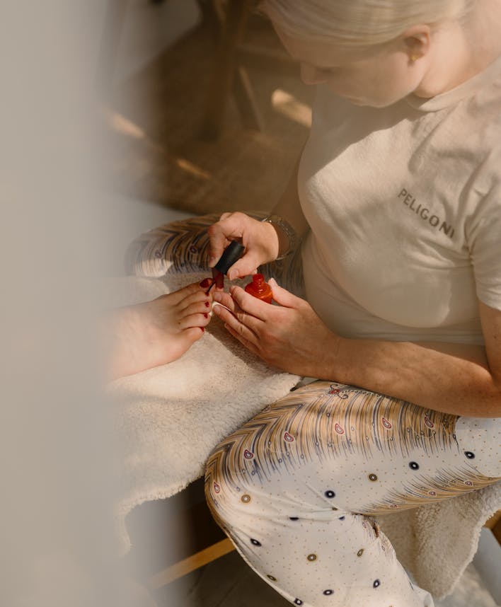 Overhead view of a nail technician applying red polish to a client's nails during a manicure session. The technician, wearing a beige 'PELIGONI' t-shirt, carefully works on the client's hand which rests on a white towel. The client wears patterned white pants with peacock feather designs and colourful dots. The image has an artistic double-exposure effect creating a dreamy, soft-focus aesthetic.