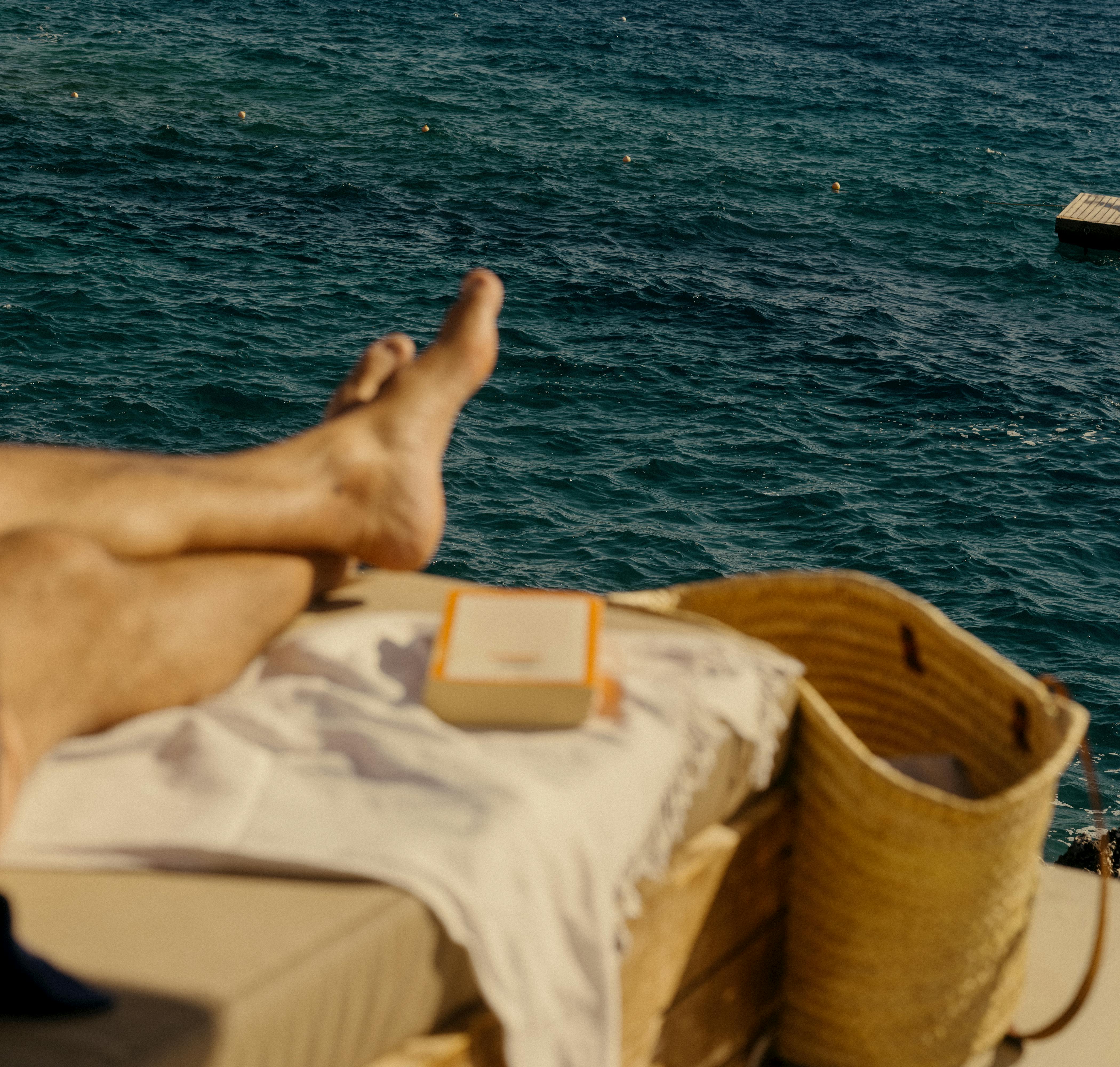 A relaxed scene by the deep blue Mediterranean Sea showing a person's leg and foot extended toward the water from a beige cushioned lounger. On the lounger sits a striped white towel, a small orange-bordered item, and a tan woven beach bag. Multiple swimmers can be seen as small dots in the water, with the horizon stretching across the frame under an overcast sky. A wooden dock edge is visible in the upper right corner.