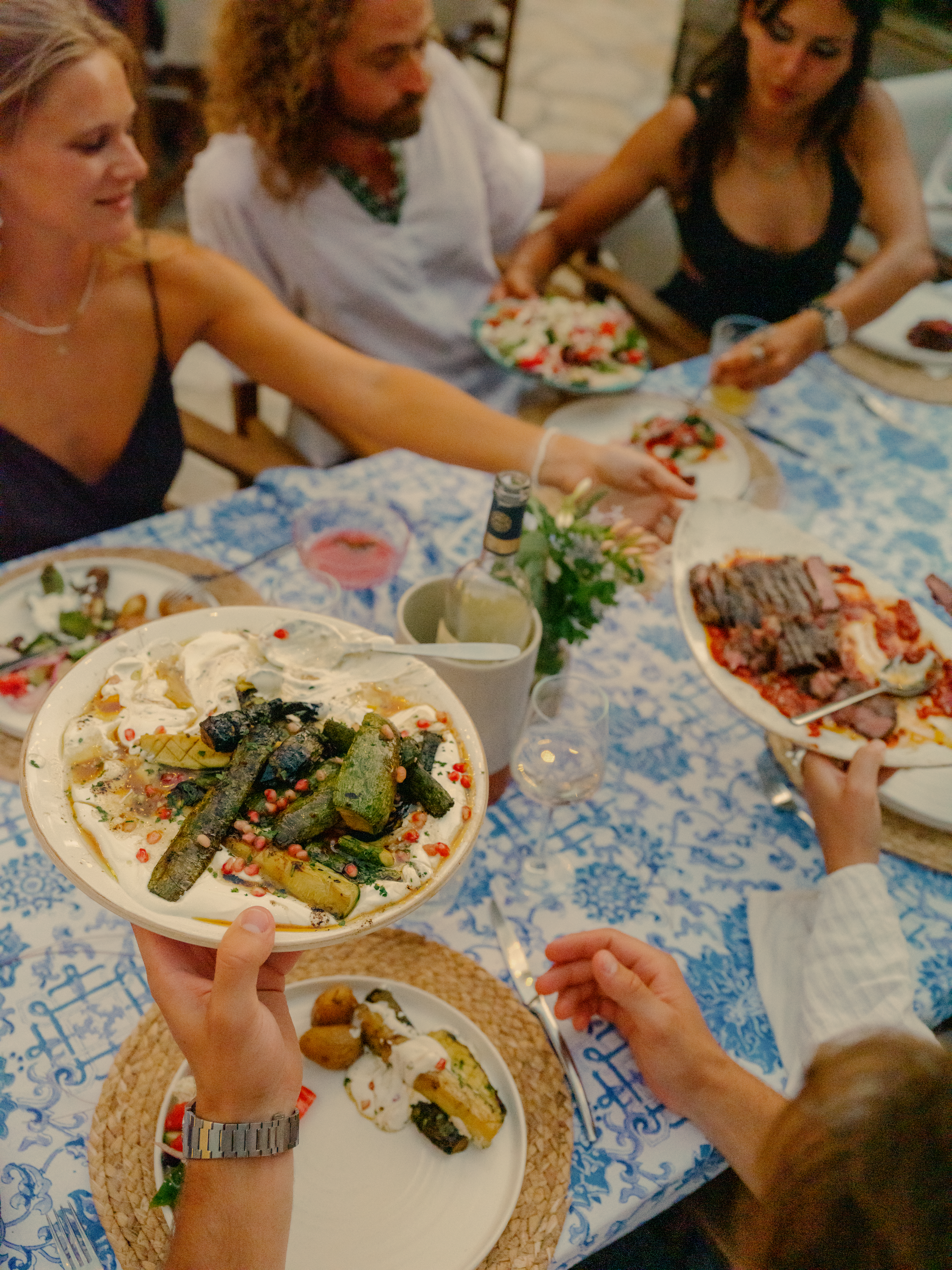 A group of people gathered around a table with a blue and white patterned tablecloth, sharing a Mediterranean-style meal family-style. The central focus is on hands holding a white plate with grilled zucchini topped with yogurt sauce, herbs, and chili flakes. The table features multiple shared dishes including grilled vegetables, salads with tomatoes, sliced meat, and small bowls of sauces. Guests in casual summer attire reach across the table to serve themselves in a warm, communal dining atmosphere.