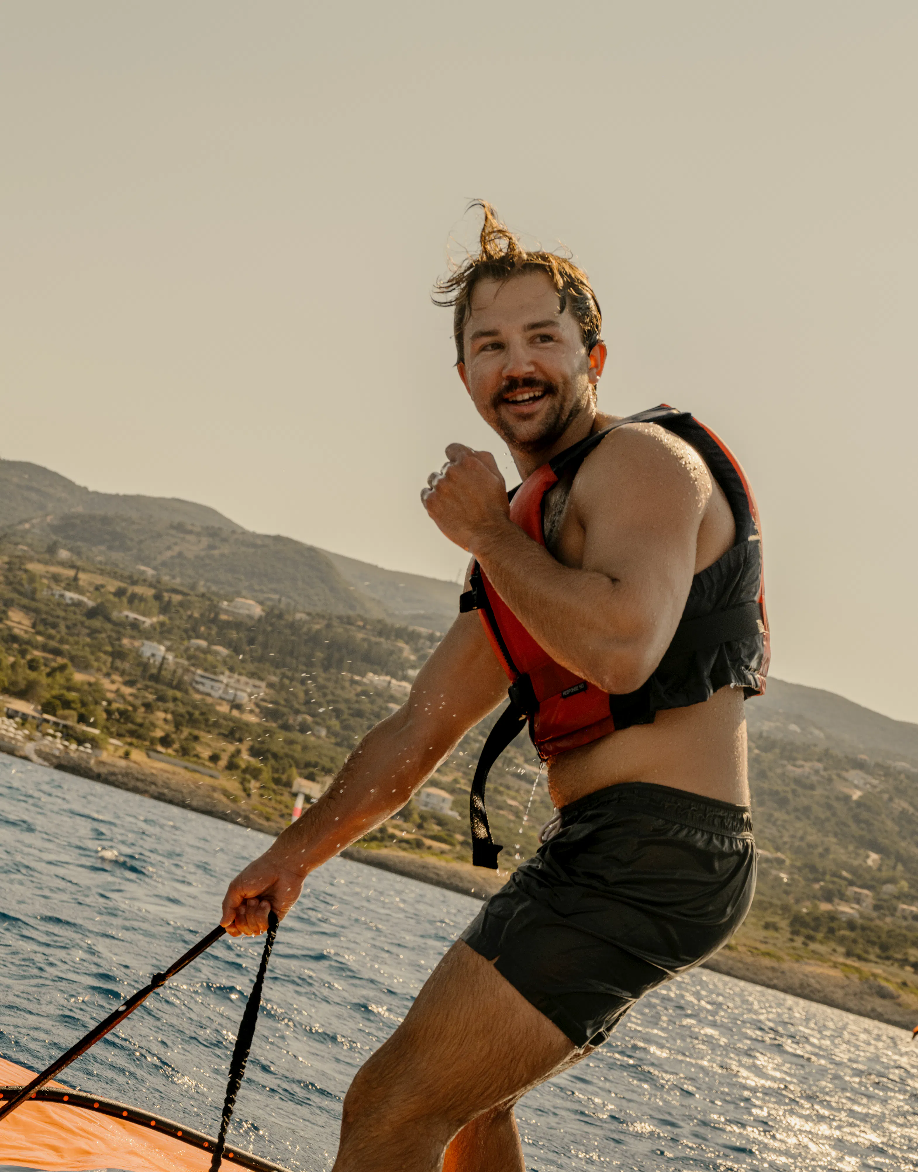A man windsurfing on sparkling water, standing on a white and blue board while holding a control bar for an orange and black wing sail. He wears black board shorts, a red and black life vest, and a yellow patterned headband, smiling at the camera. The background shows a hillside coastline with scattered vegetation under warm lighting conditions.