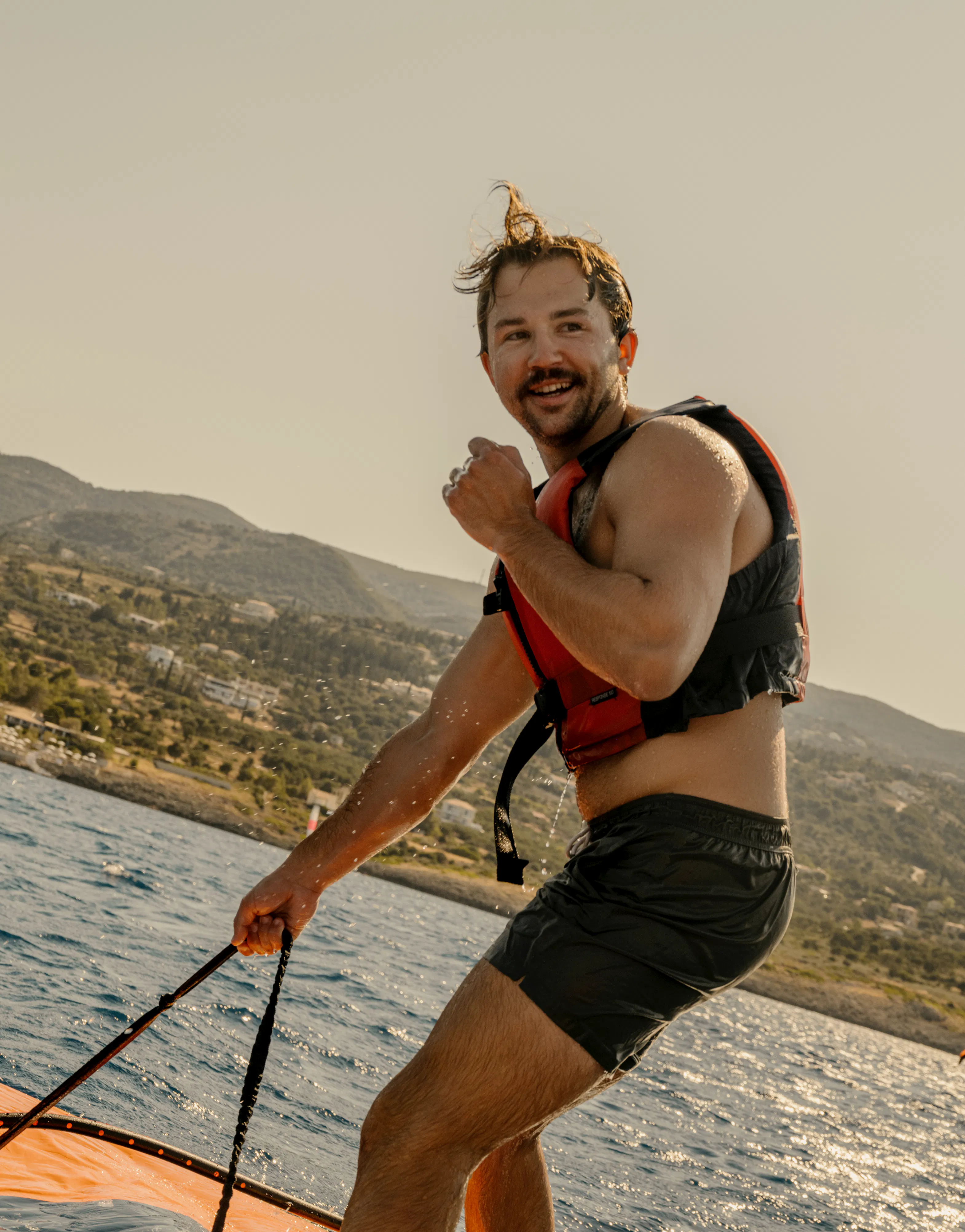 A man windsurfing on sparkling water, standing on a white and blue board while holding a control bar for an orange and black wing sail. He wears black board shorts, a red and black life vest, and a yellow patterned headband, smiling at the camera. The background shows a hillside coastline with scattered vegetation under warm lighting conditions.