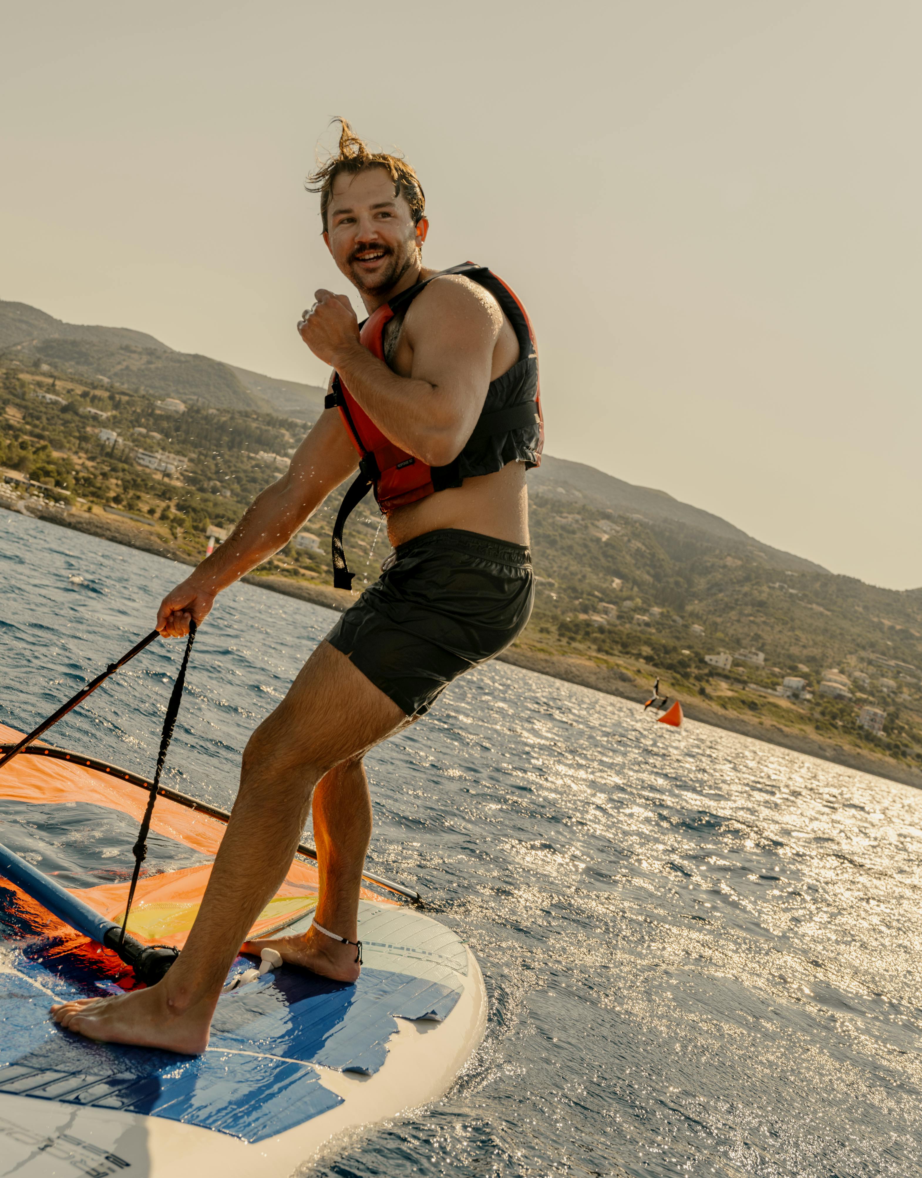 A man windsurfing on sparkling water, standing on a white and blue board while holding a control bar for an orange and black wing sail. He wears black board shorts, a red and black life vest, and a yellow patterned headband, smiling at the camera. The background shows a hillside coastline with scattered vegetation under warm lighting conditions.