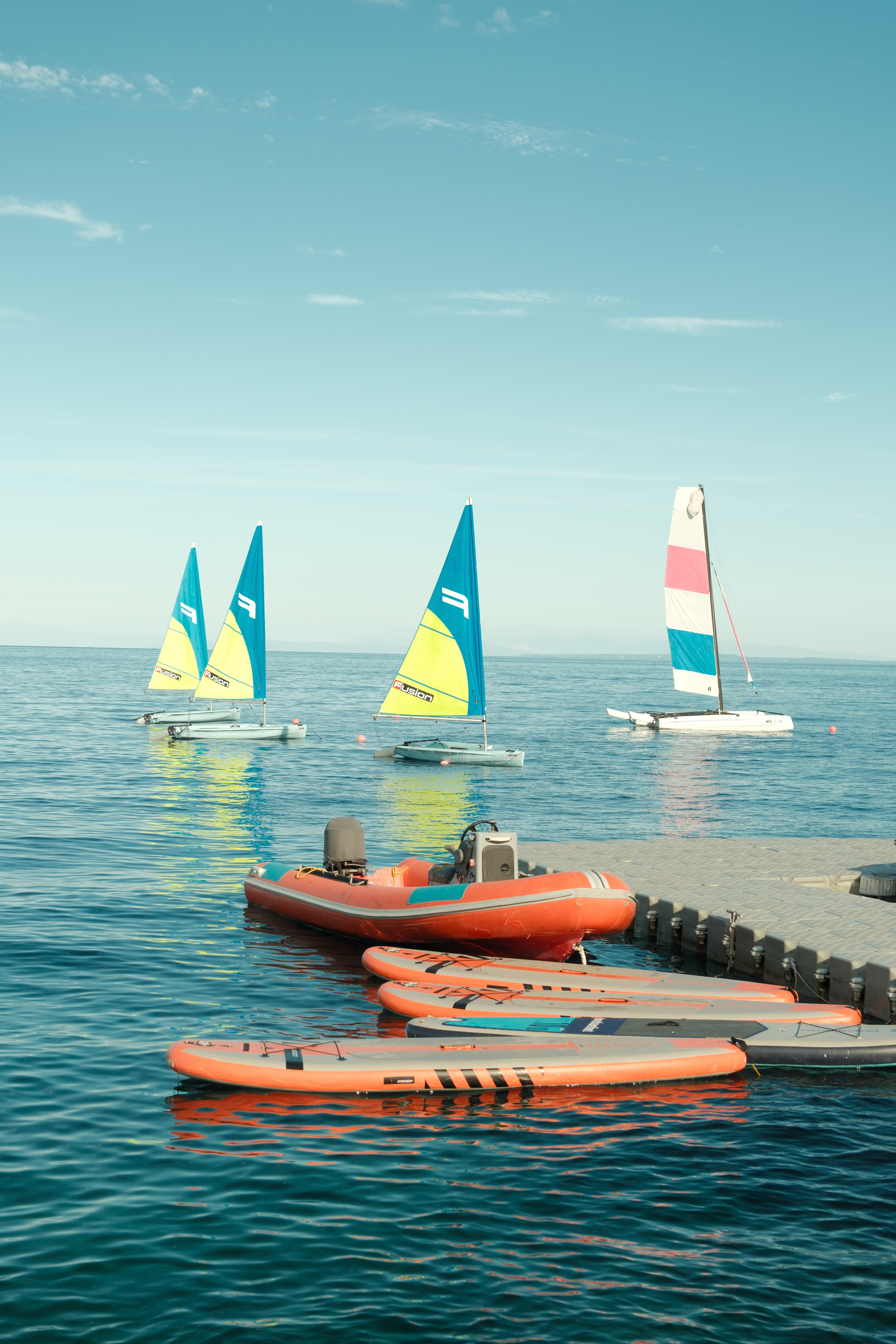 A serene water sports scene showing three small catamarans with colorful sails (turquoise with yellow accents, and white with pink/blue stripes) anchored in calm blue water. In the foreground, an orange inflatable boat and several orange paddleboards or hydrofoils are lined up at the water's edge. The clear sky and tranquil water create a perfect setting for water activities.
