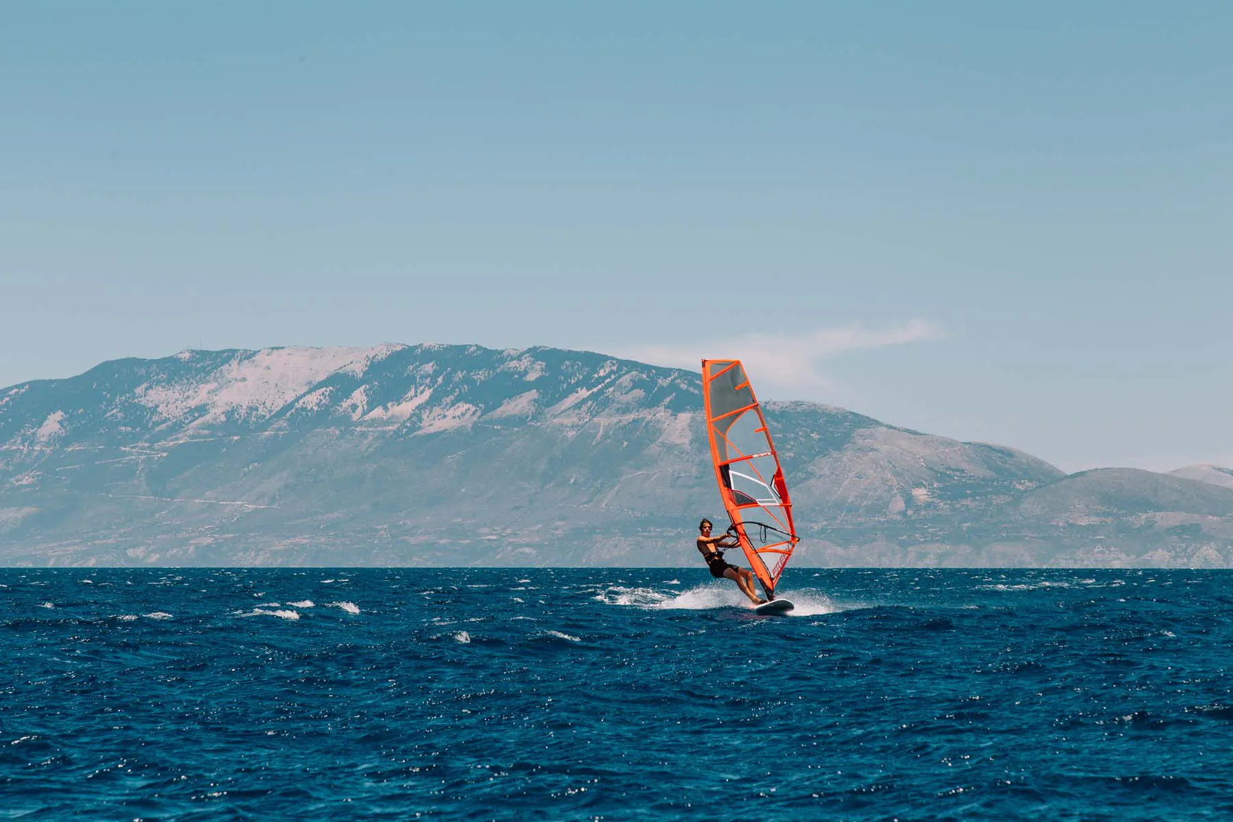 A windsurfer riding choppy blue waters with a bright orange and white sail, set against a dramatic backdrop of mountains with snow-dusted peaks. White spray kicks up around the board as the surfer navigates the waves under a pale blue sky with wispy clouds.