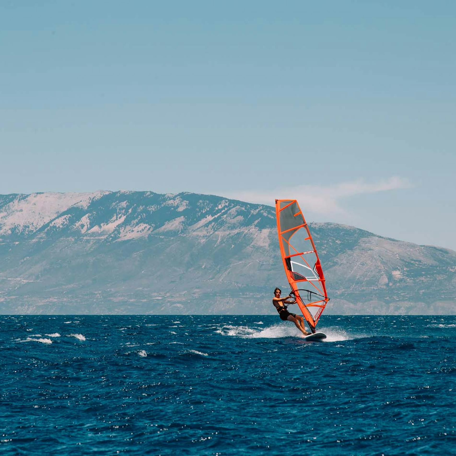 A windsurfer riding choppy blue waters with a bright orange and white sail, set against a dramatic backdrop of mountains with snow-dusted peaks. White spray kicks up around the board as the surfer navigates the waves under a pale blue sky with wispy clouds.
