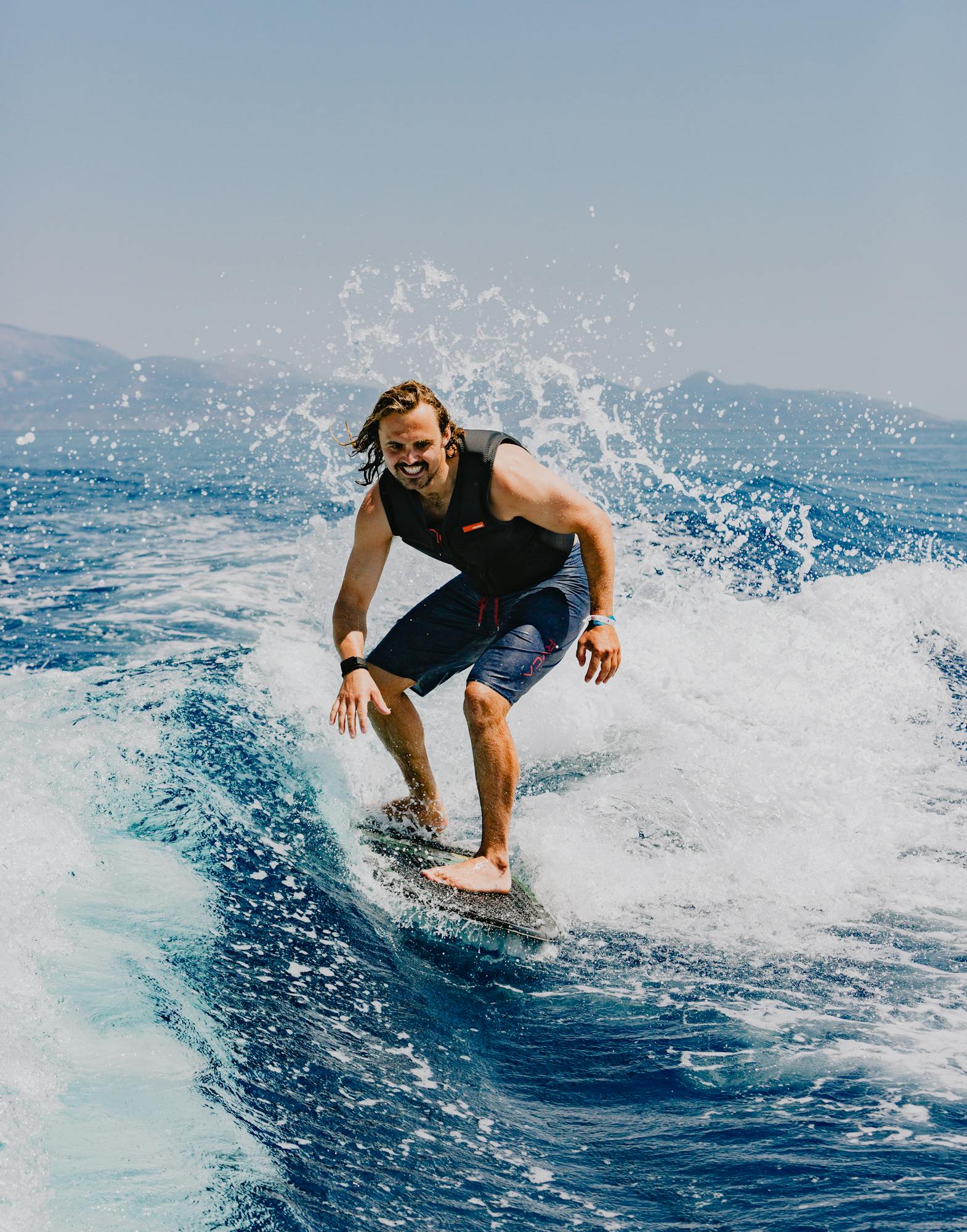 A smiling surfer riding a wave on a blue surfboard, wearing a black wetsuit vest and blue board shorts. Water sprays dramatically around them as they navigate the white foam on the deep blue ocean water. Mountains are visible in the hazy background under a clear sky.