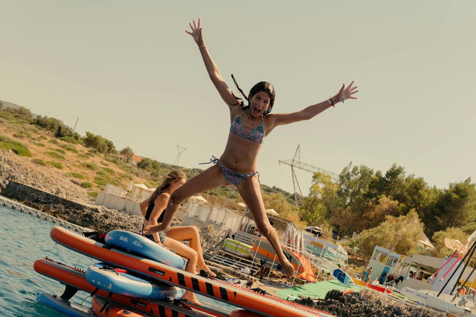 A girl in a patterned bikini jumps joyfully with arms spread wide from a floating inflatable obstacle course, while others lounge on the structure in the turquoise water. The coastal setting shows a hillside beach area with buildings, greenery, and equipment in the background. Someone swims in the water below the jump platform, which features orange and blue striped bars.