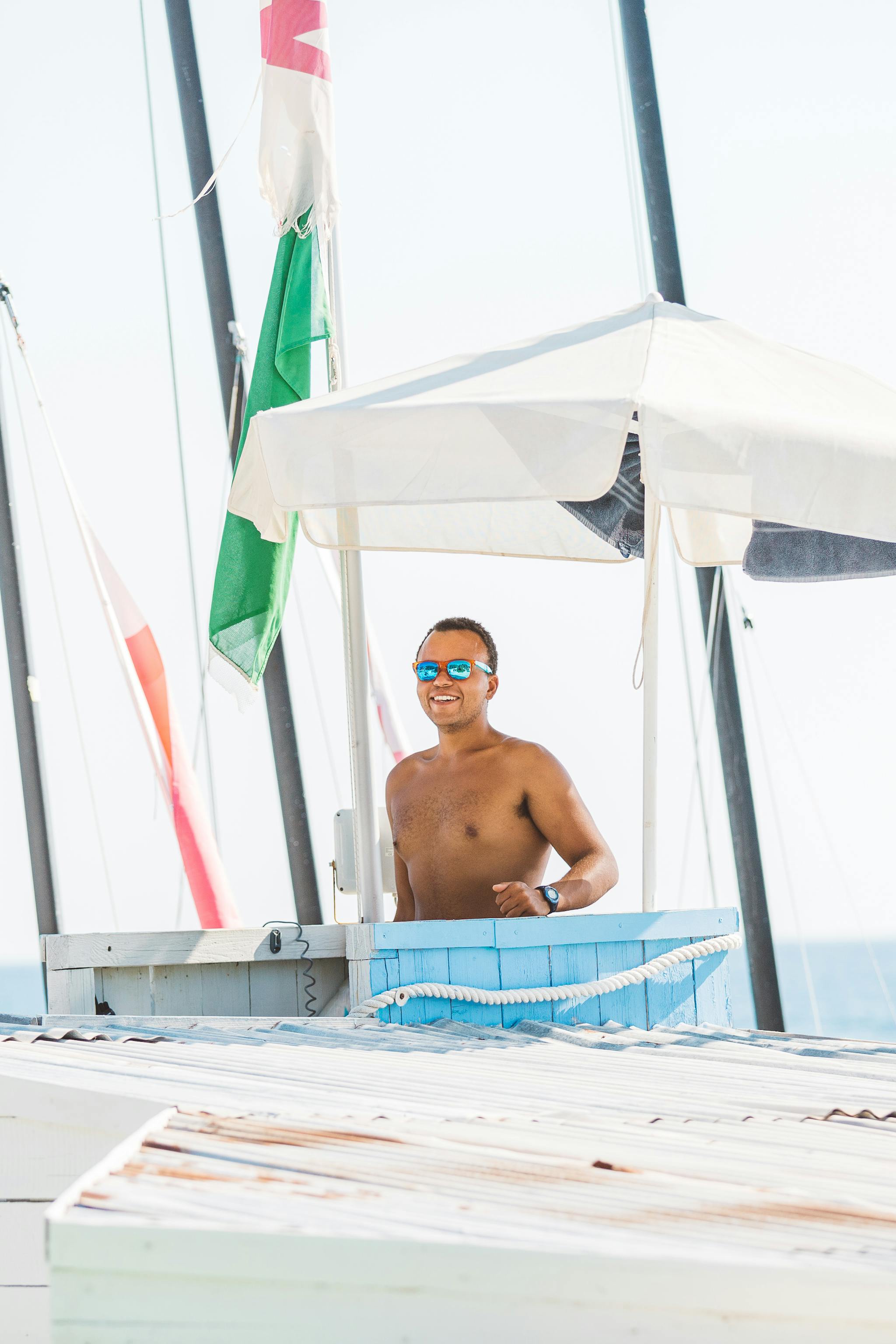 A shirtless man wearing blue mirrored sunglasses smiles from inside a white beach kiosk or towel station with an open-air design. The structure has a white canopy roof, blue towels visible on the counter, and sailing masts with rigging visible in the background against a bright, overcast sky. The scene appears to be at a waterfront or marina location.