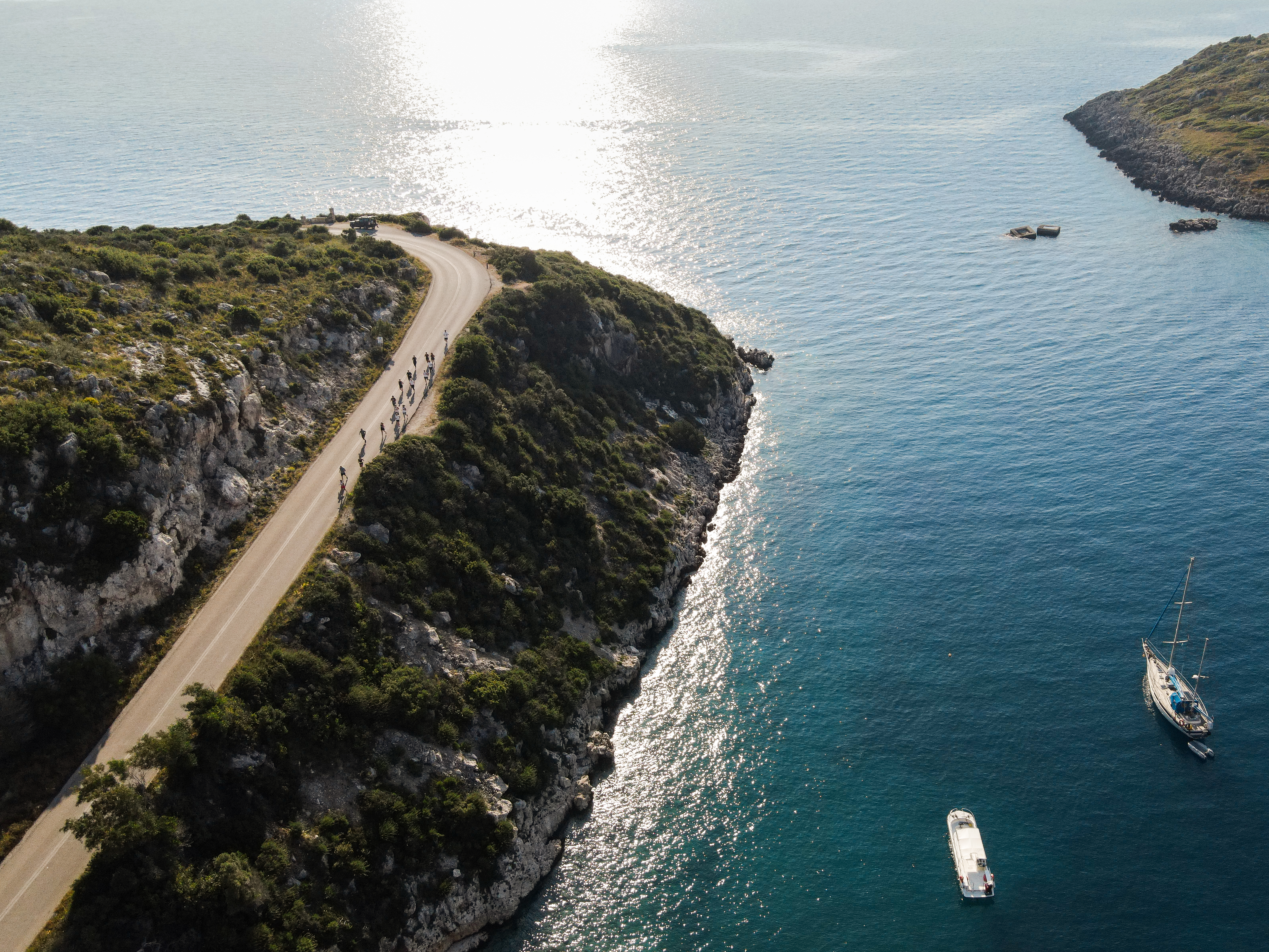Aerial view of a scenic coastal road winding along dramatic limestone cliffs covered in Mediterranean vegetation. A group of cyclists rides along the curved asphalt road that hugs the rocky coastline. Below, turquoise waters shimmer in the sunlight, with two sailboats anchored in the calm bay. The contrast between the rugged white cliffs, lush greenery, and deep blue sea creates a stunning Mediterranean landscape.