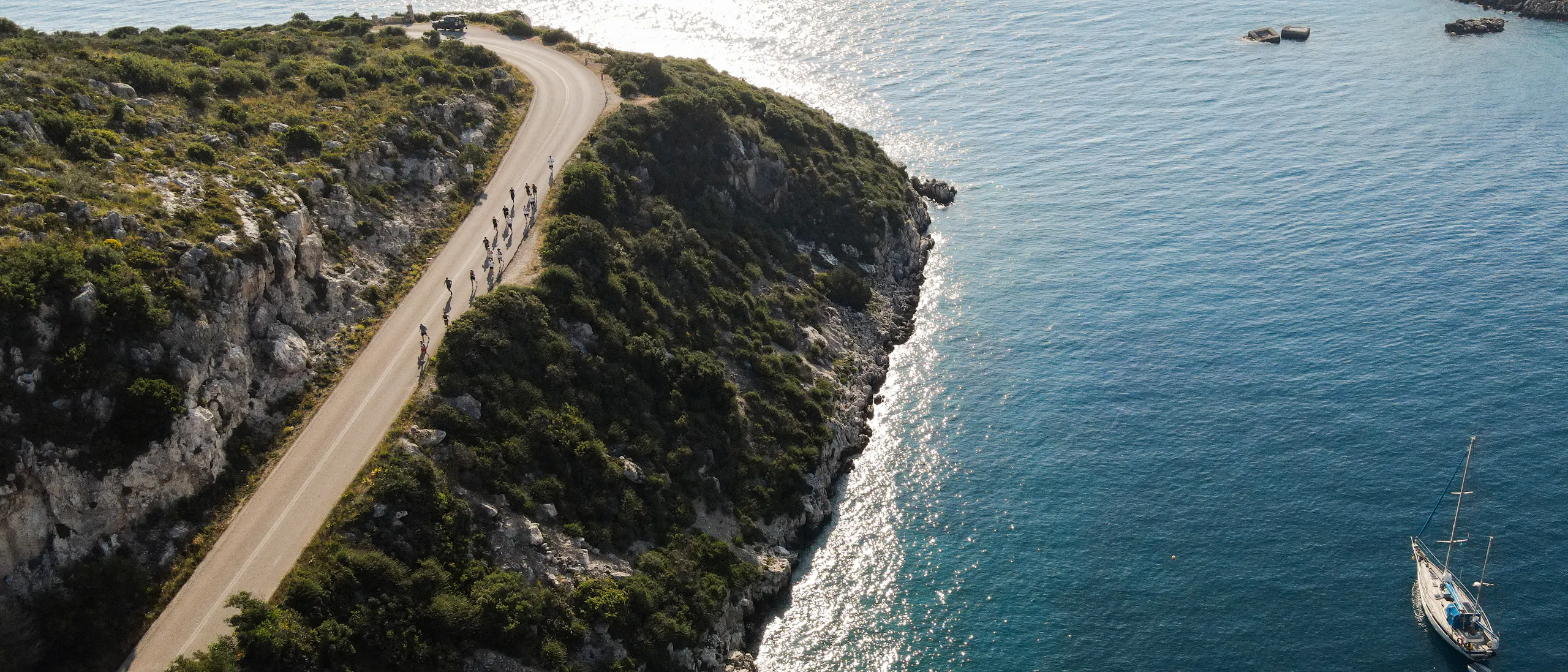 Aerial view of a scenic coastal road winding along dramatic limestone cliffs covered in Mediterranean vegetation. A group of cyclists rides along the curved asphalt road that hugs the rocky coastline. Below, turquoise waters shimmer in the sunlight, with two sailboats anchored in the calm bay. The contrast between the rugged white cliffs, lush greenery, and deep blue sea creates a stunning Mediterranean landscape.