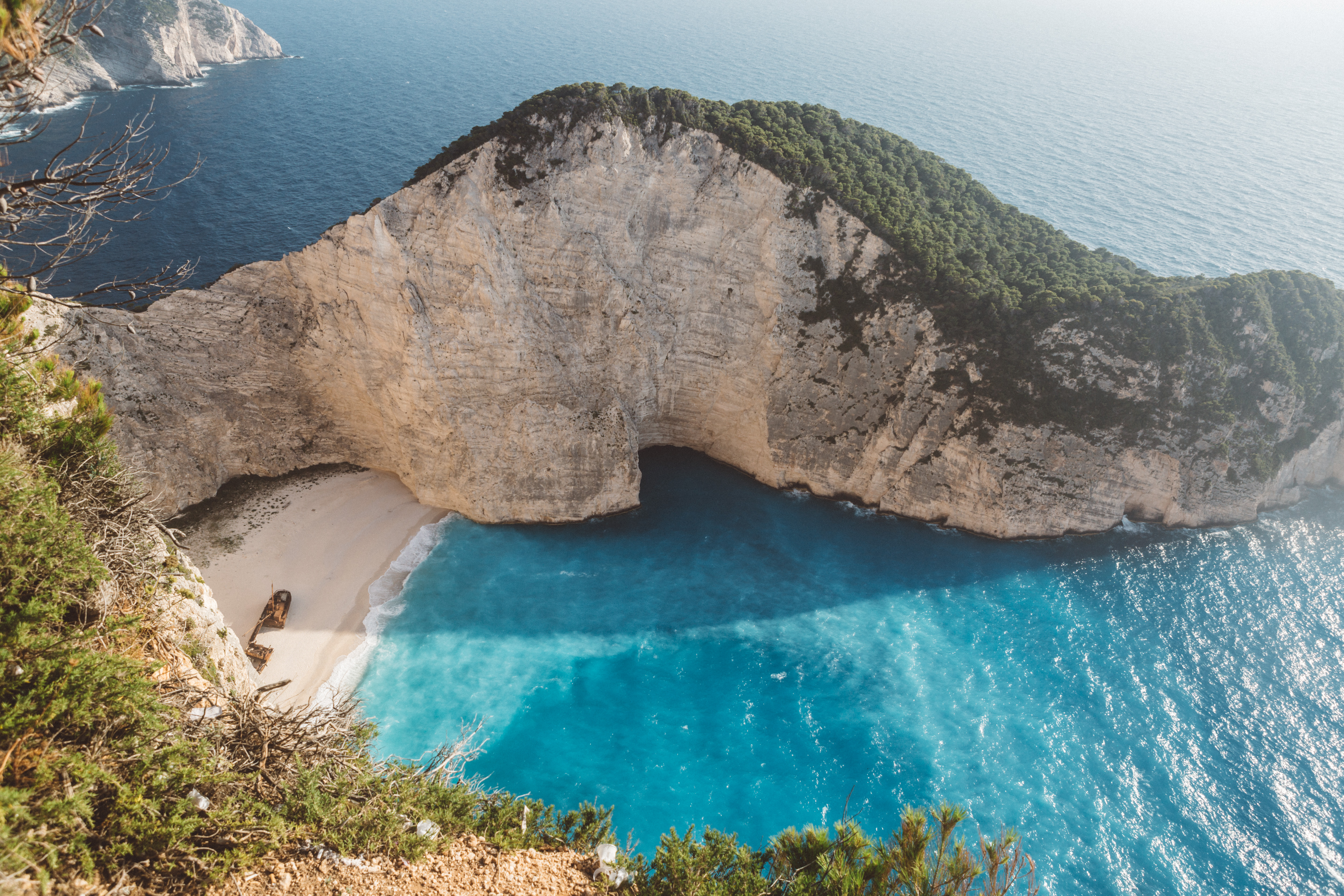 Aerial view of the famous Navagio Beach (Shipwreck Beach) in Zakynthos, Greece, showing dramatic white limestone cliffs towering over a secluded cove with turquoise waters. A rusted shipwreck rests on the pristine white sand beach. Lush green vegetation frames the top and sides of the cliff edges, while the deep blue Mediterranean Sea extends to the horizon where another rocky headland is visible in the distance.