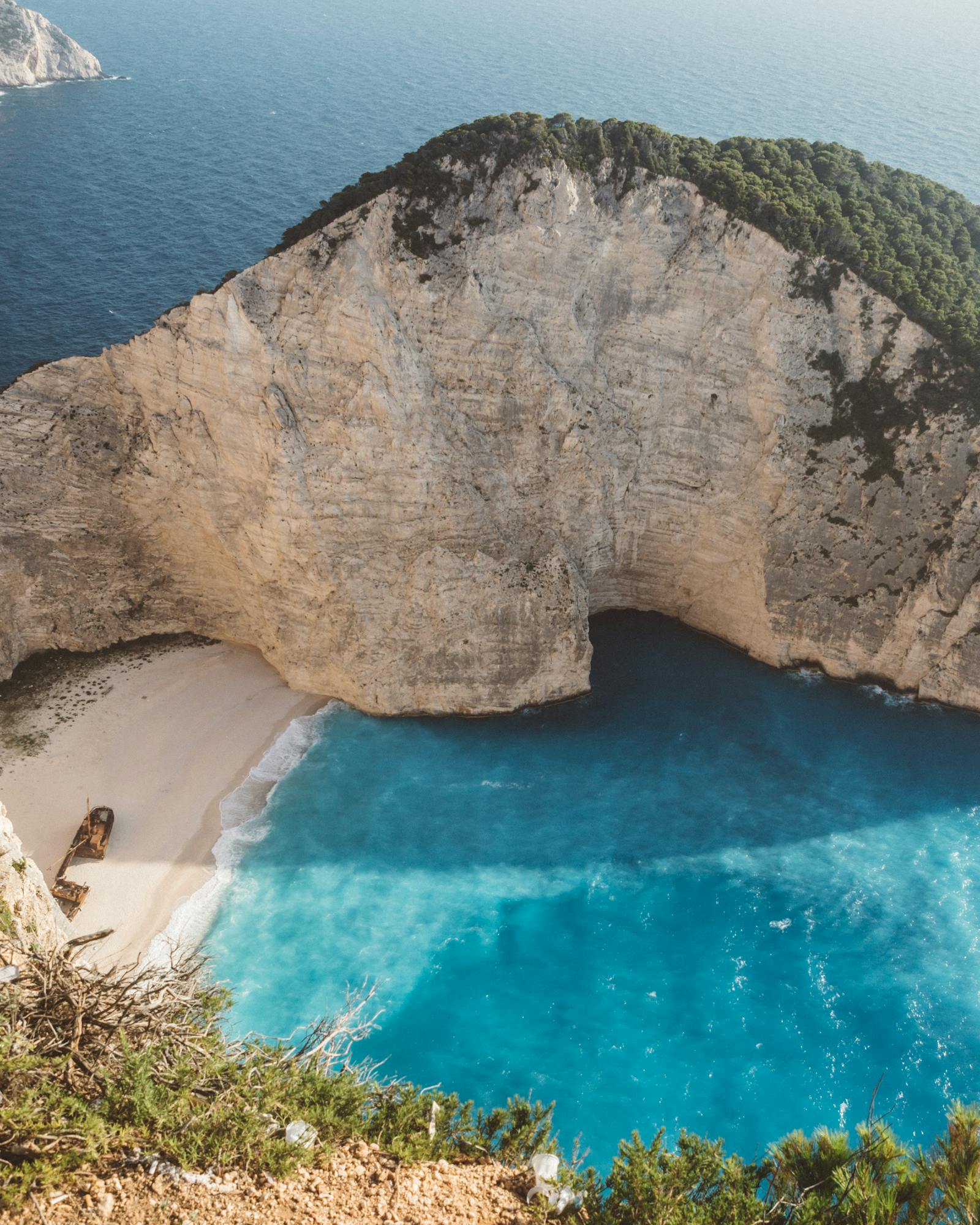 Aerial view of the famous Navagio Beach (Shipwreck Beach) in Zakynthos, Greece, showing dramatic white limestone cliffs towering over a secluded cove with turquoise waters. A rusted shipwreck rests on the pristine white sand beach. Lush green vegetation frames the top and sides of the cliff edges, while the deep blue Mediterranean Sea extends to the horizon where another rocky headland is visible in the distance.