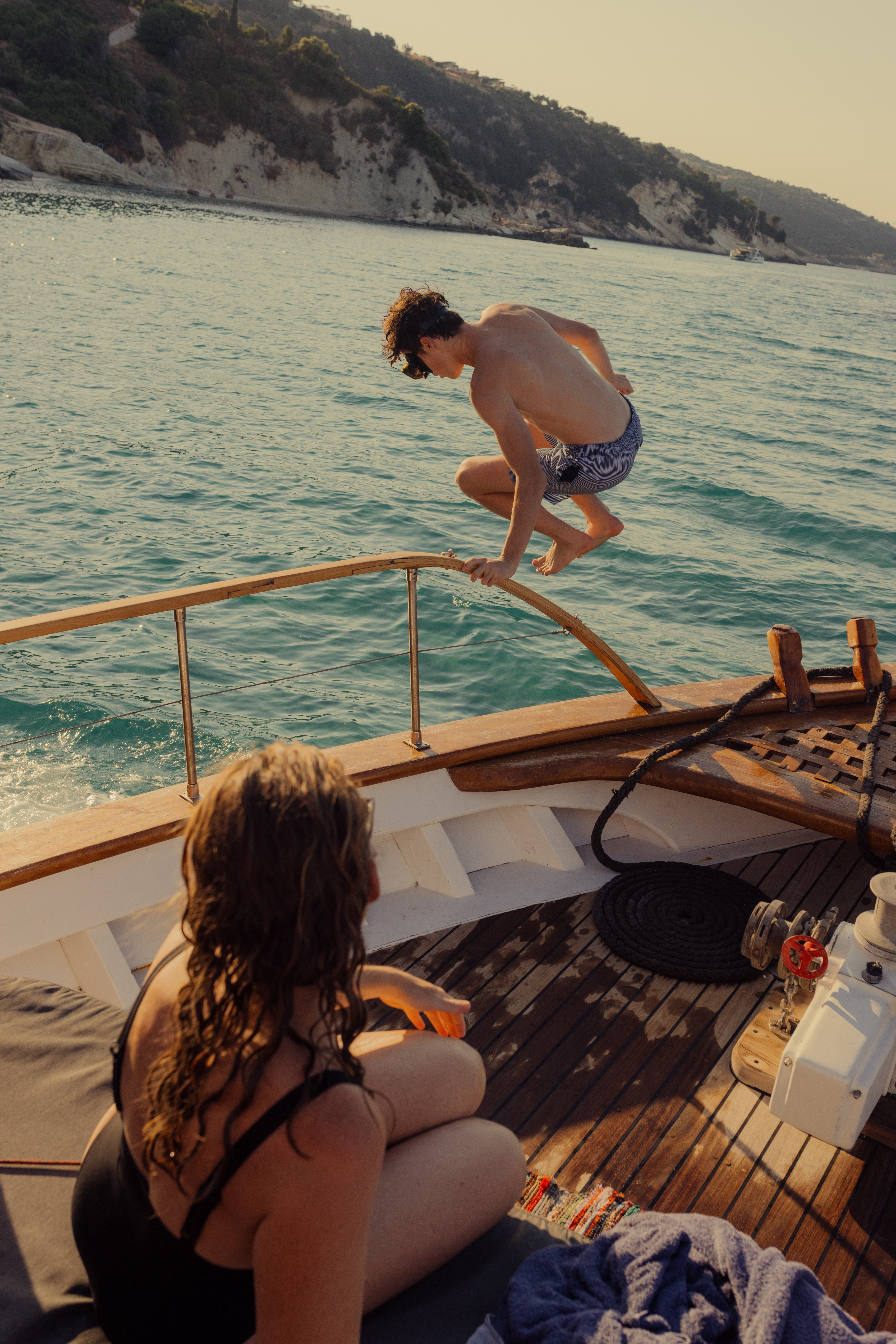 A young person mid-jump from a wooden boat deck into turquoise coastal waters, with white cliffs and vegetation visible in the background. Another person with long wet hair watches from the boat's seating area. The boat features a classic wooden deck with teak flooring, white cushioned seating, metal railings, and nautical equipment. The scene captures a summer day of coastal leisure and water activities.