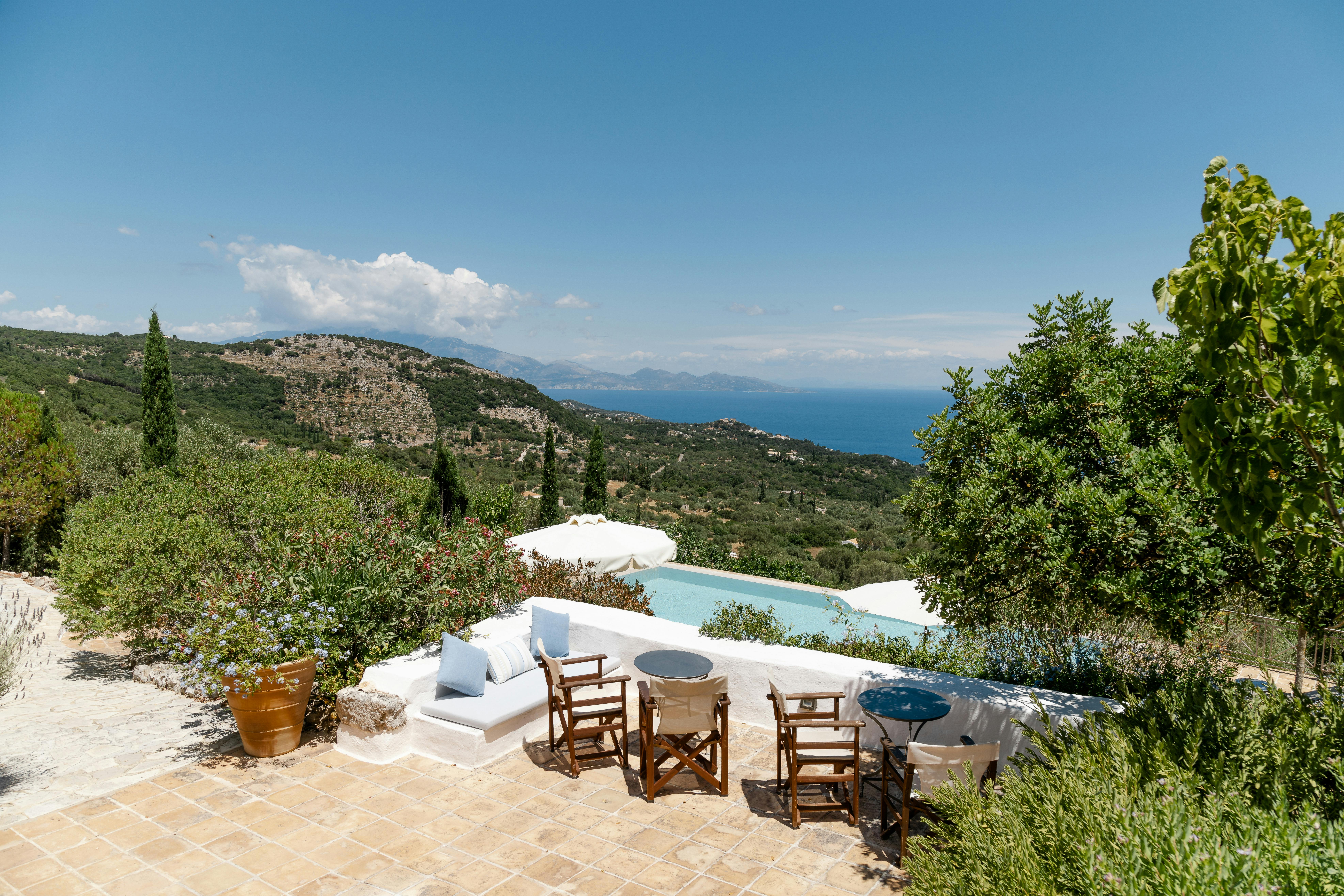 White curved seating area and folding director's chairs around rustic table beside infinity pool overlooking hills and sea. Terracotta tiles and Mediterranean landscape complete the scenic terrace.