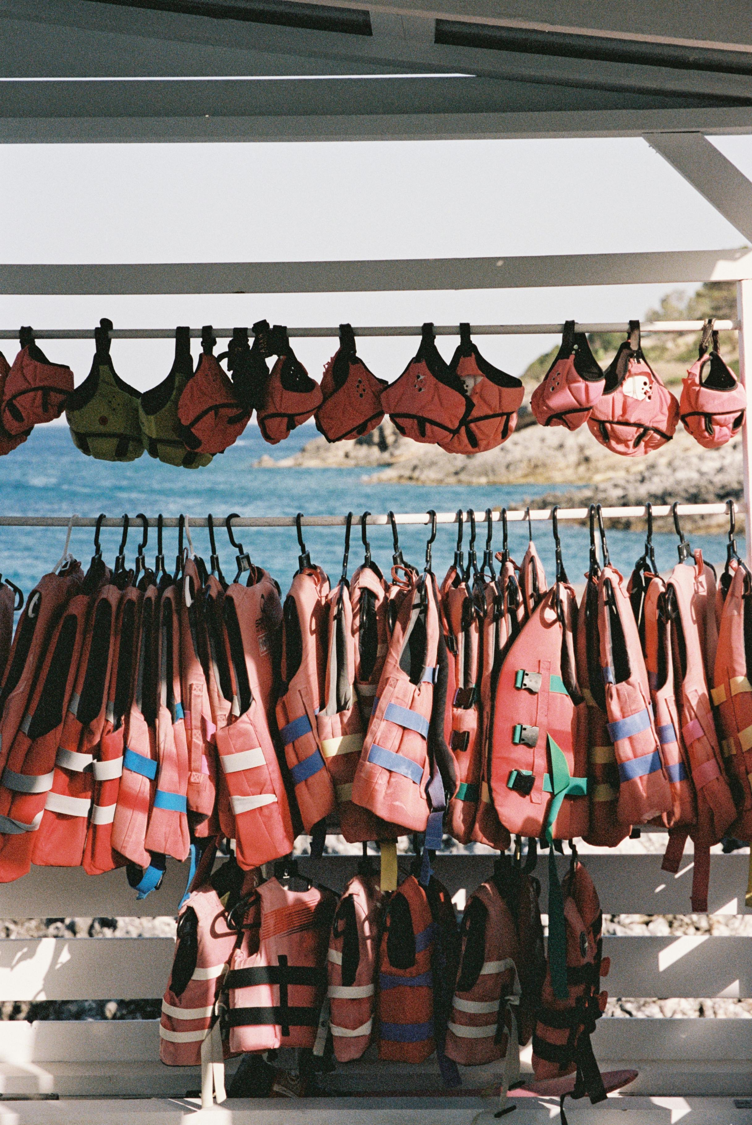 Rows of orange life jackets hang organised on racks under a white canopy or overhang. The life jackets are arranged on multiple levels - some hanging upside down on the top row, and others hanging normally on lower rows. The jackets feature black straps and buckles, with some showing reflective striping. Bright sunlight filters through the storage area, and water or a marine environment is visible in the background.
