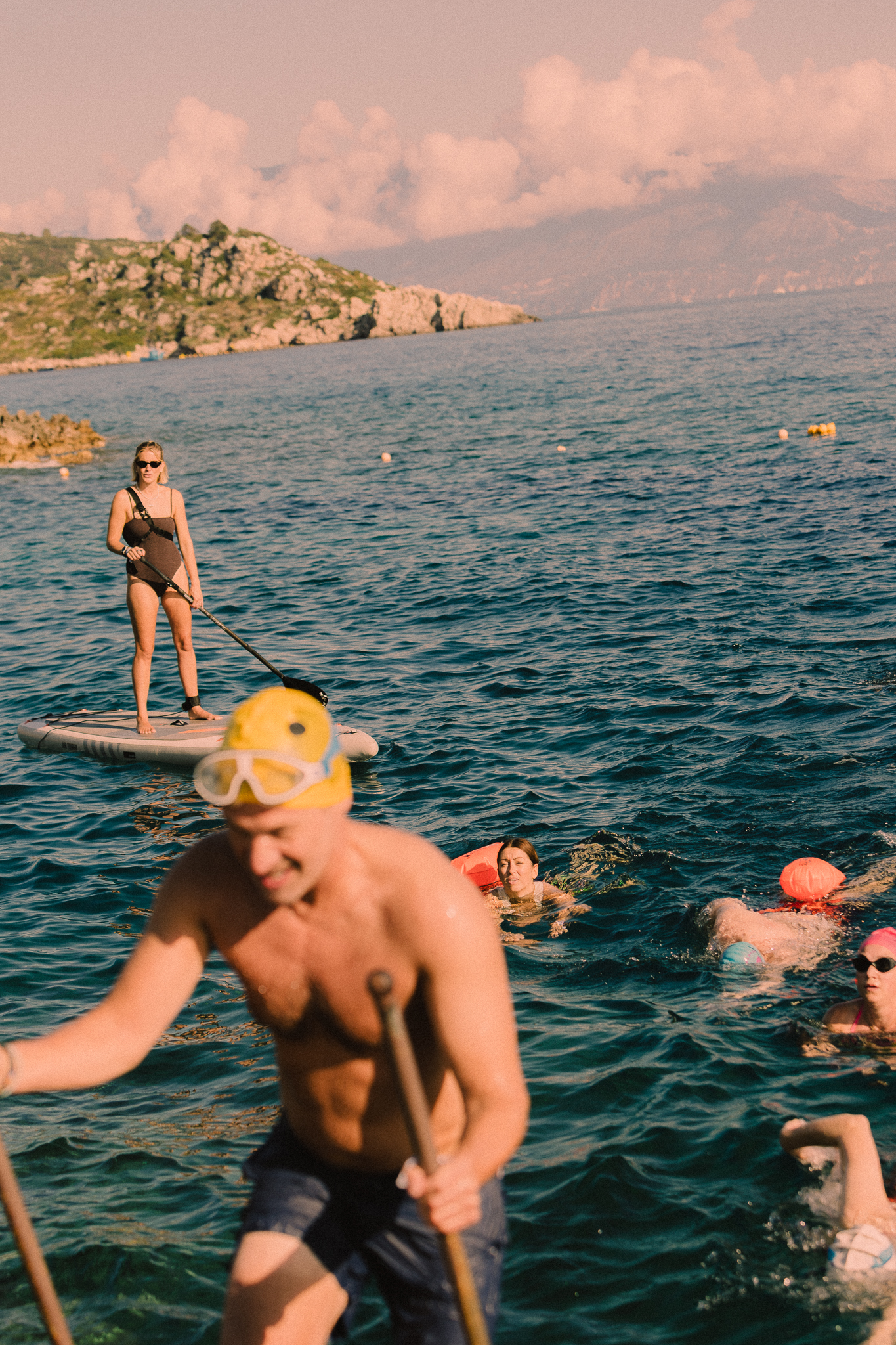 A group enjoying a golden-hour swim in deep blue water. In the foreground, a person wearing a yellow snorkel mask is partially visible and out of focus. Behind them, a woman in a black swimsuit and sunglasses stands on a paddleboard holding a paddle, while several people swim in the water wearing orange flotation devices and snorkel gear. In the background, rocky coastal hills covered with scrub vegetation rise from the water under dramatic pink-tinged clouds at sunset.