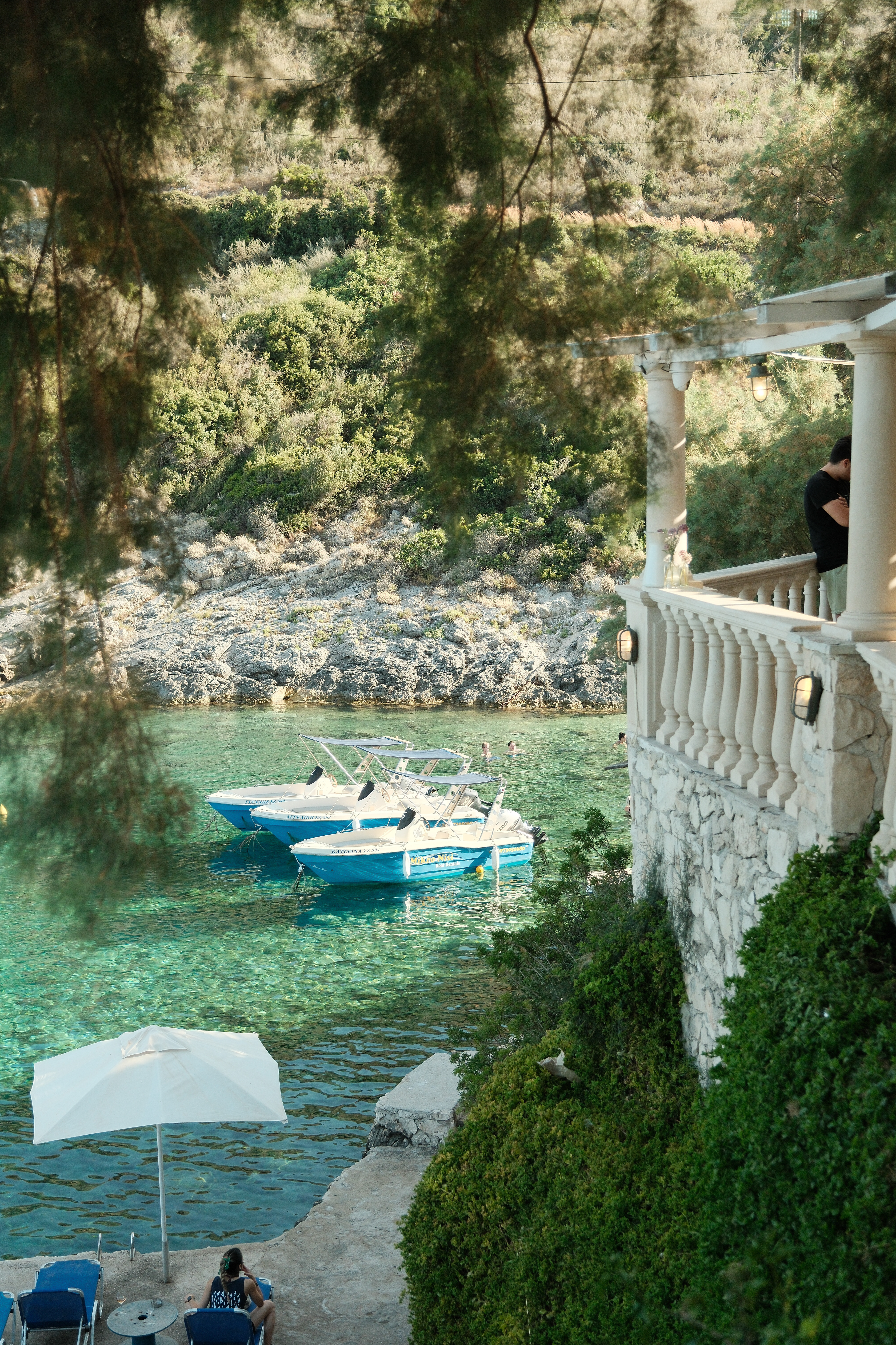 A serene cove with crystal-clear turquoise water, viewed from a white ornate balcony with classical balustrades. Two small boats are moored in the protected inlet, surrounded by a rocky shoreline covered in dense green vegetation and trees. Below, a narrow concrete dock extends into the water where people can be seen relaxing under a white umbrella. The hillside behind is rugged and covered with Mediterranean scrub and pine trees, creating a peaceful, secluded atmosphere.