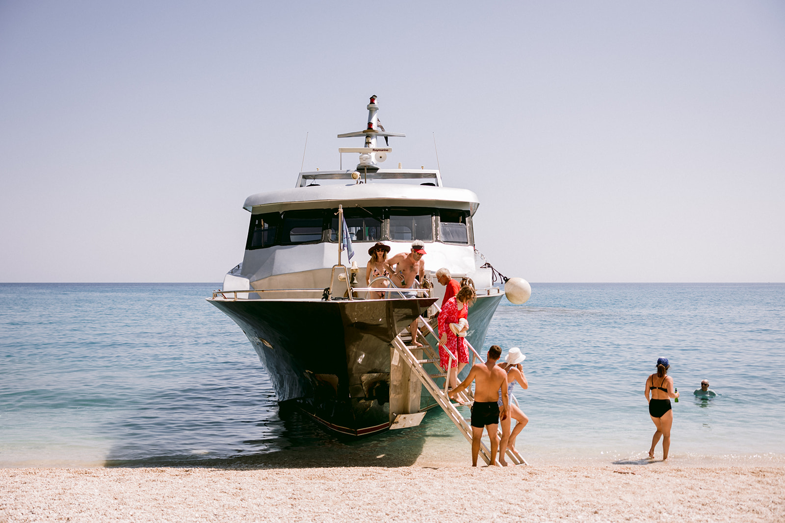 A white and dark green luxury motor yacht beached on a white pebble shore in crystal-clear turquoise water. Guests disembark down a gangway, including a woman in a vibrant red floral dress. Several people are visible on the yacht's deck under the flybridge, while others walk along the beach in swimwear - some heading toward the water. A person stands atop the flybridge against the pale blue sky. Swimmers can be seen in the calm, shallow water to the right. The scene captures a leisurely Mediterranean summer day with the yacht serving as transport for beachgoers to this pristine coastal spot.