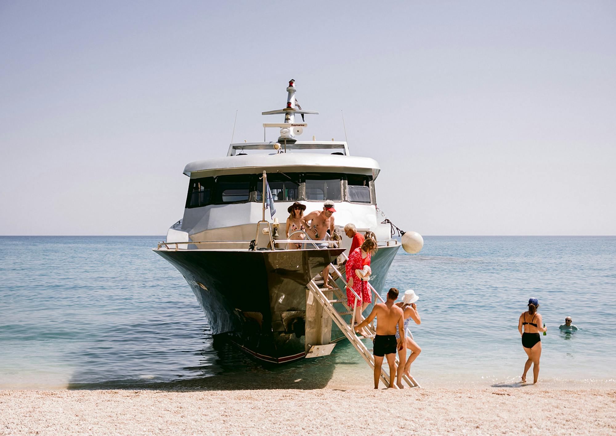 A white and dark green luxury motor yacht beached on a white pebble shore in crystal-clear turquoise water. Guests disembark down a gangway, including a woman in a vibrant red floral dress. Several people are visible on the yacht's deck under the flybridge, while others walk along the beach in swimwear - some heading toward the water. A person stands atop the flybridge against the pale blue sky. Swimmers can be seen in the calm, shallow water to the right. The scene captures a leisurely Mediterranean summer day with the yacht serving as transport for beachgoers to this pristine coastal spot.