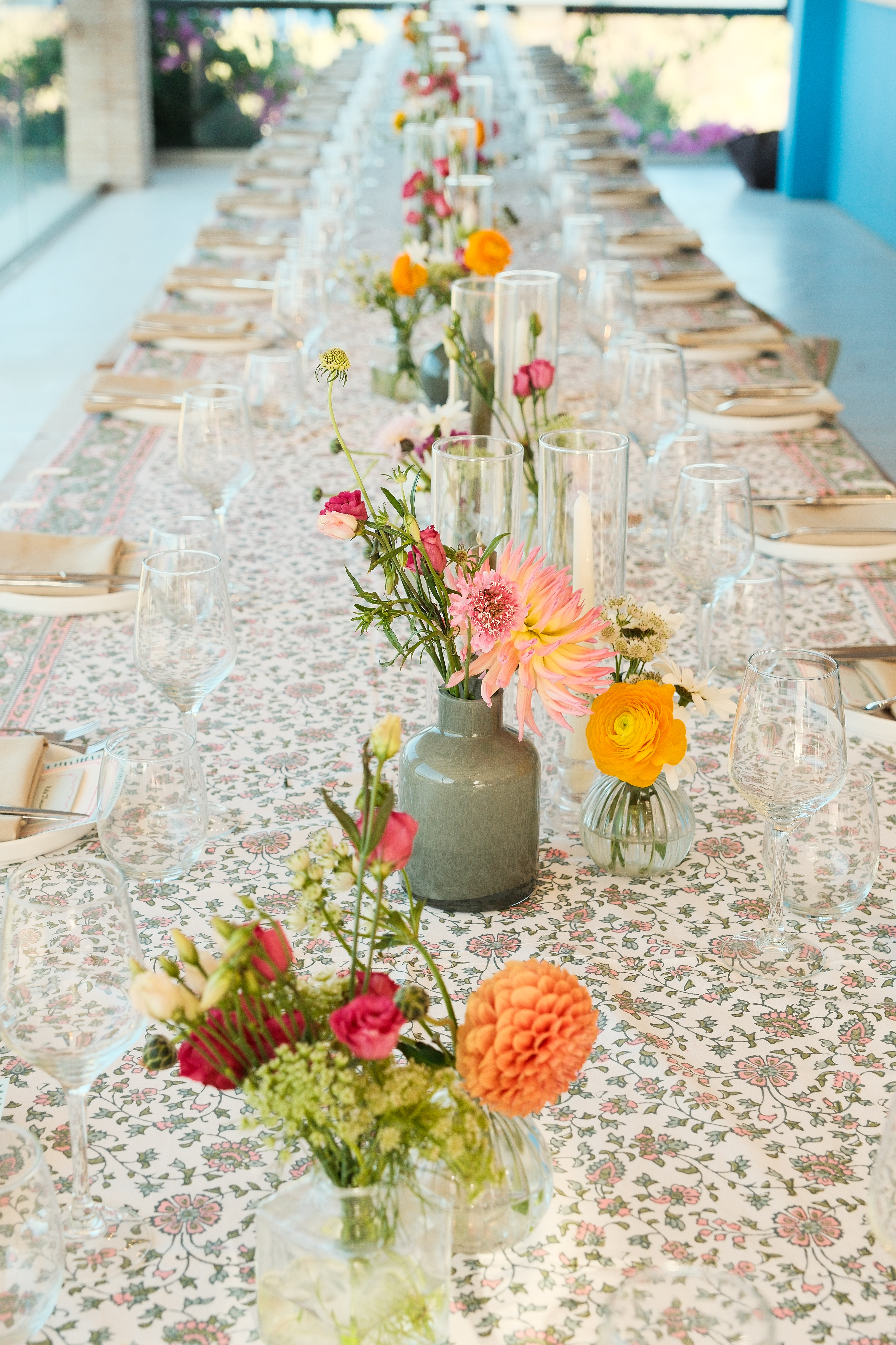 A long dining table set for an elegant outdoor event, photographed lengthwise to show the repeating pattern of place settings stretching into soft focus. The table is covered with a delicate floral-patterned tablecloth in soft pastels. Down the center, a series of small floral arrangements in varied vases feature cheerful blooms - coral dahlias, bright yellow ranunculus, pink spray roses, and wildflowers in muted ceramic vessels and clear glass bottles. Each place setting includes natural woven chargers, folded napkins, and multiple wine glasses. Tall cylinder vases with single stems create vertical interest between the lower arrangements. The bright, airy setting with white walls and colorful flowers visible in the blurred background creates a fresh, summery Mediterranean garden party atmosphere.