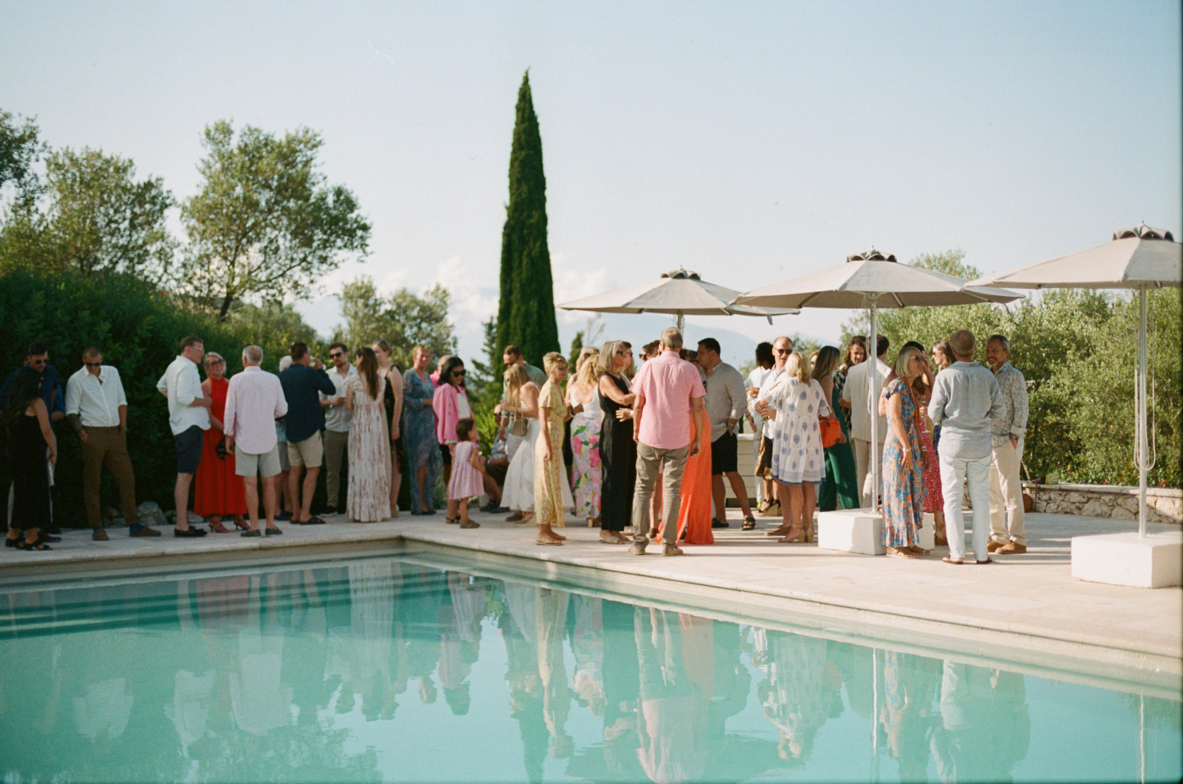 An elegant outdoor cocktail reception beside a pristine turquoise swimming pool on a warm afternoon. Guests in smart casual and semi-formal summer attire mingle on the pale stone pool deck, their figures and the tall beige market umbrellas reflected in the still water. The crowd includes adults and children wearing colorful sundresses, linen shirts, and light trousers in shades of pink, coral, white, and floral patterns. A distinctive tall Italian cypress tree stands as a vertical accent in the center background, flanked by olive trees and lush Mediterranean vegetation. The refined, relaxed atmosphere and natural setting create a quintessentially Mediterranean garden party scene under soft afternoon light.