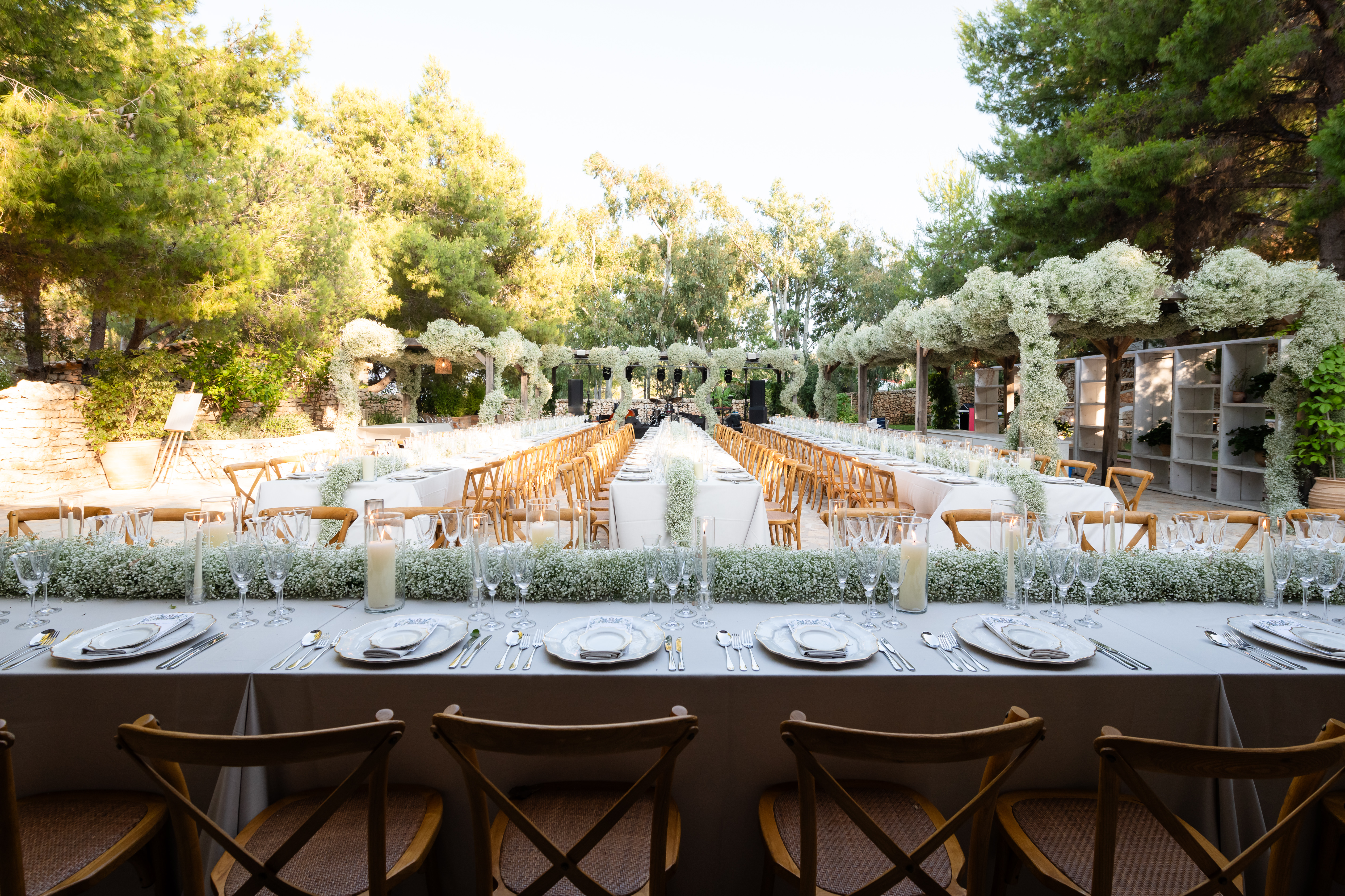 Elegant outdoor wedding reception setup with multiple long tables set with white tablecloths and place settings. Wooden cross-back chairs line the tables, which feature lush white floral runners down the middle. The venue is set in a garden courtyard surrounded by mature trees and stone buildings with an archway covered in white flowers in the background. Golden hour sunlight illuminates the romantic Mediterranean-style setting.