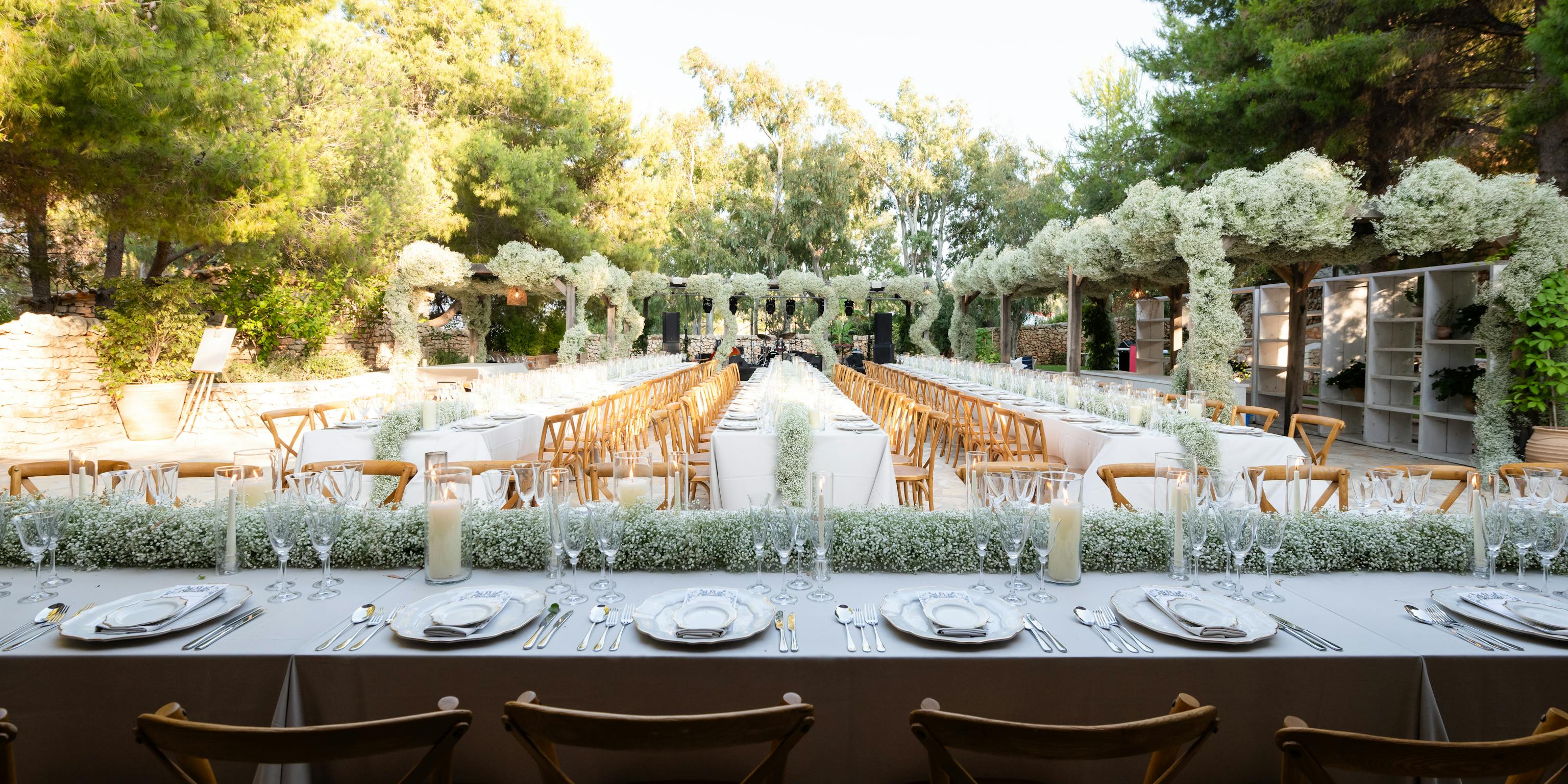 Elegant outdoor wedding reception setup with multiple long tables set with white tablecloths and place settings. Wooden cross-back chairs line the tables, which feature lush white floral runners down the middle. The venue is set in a garden courtyard surrounded by mature trees and stone buildings with an archway covered in white flowers in the background. Golden hour sunlight illuminates the romantic Mediterranean-style setting.