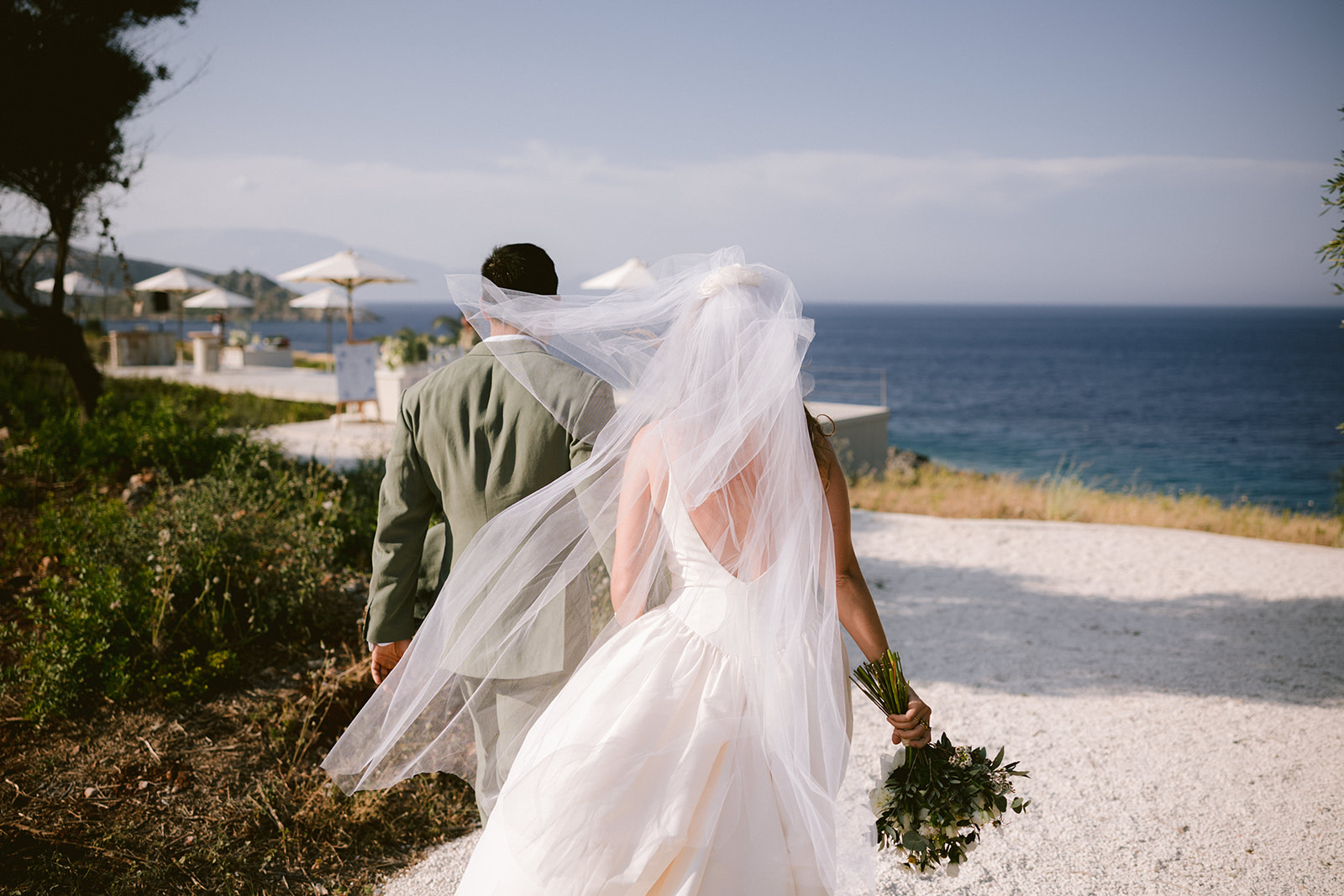 A newlywed couple photographed from behind as they walk away from their ceremony site, with the bride's voluminous white tulle veil caught dramatically in the wind, billowing across both figures. The groom wears an olive green suit while the bride in her white gown carries a lush green bouquet. They walk along a white stone path overlooking the deep blue Mediterranean Sea. In the distance, cream-coloured market umbrellas and wedding venue areas are visible on the clifftop, with misty blue hills and coastline stretching across the horizon. The romantic, windswept moment captures the joy of their seaside celebration.
