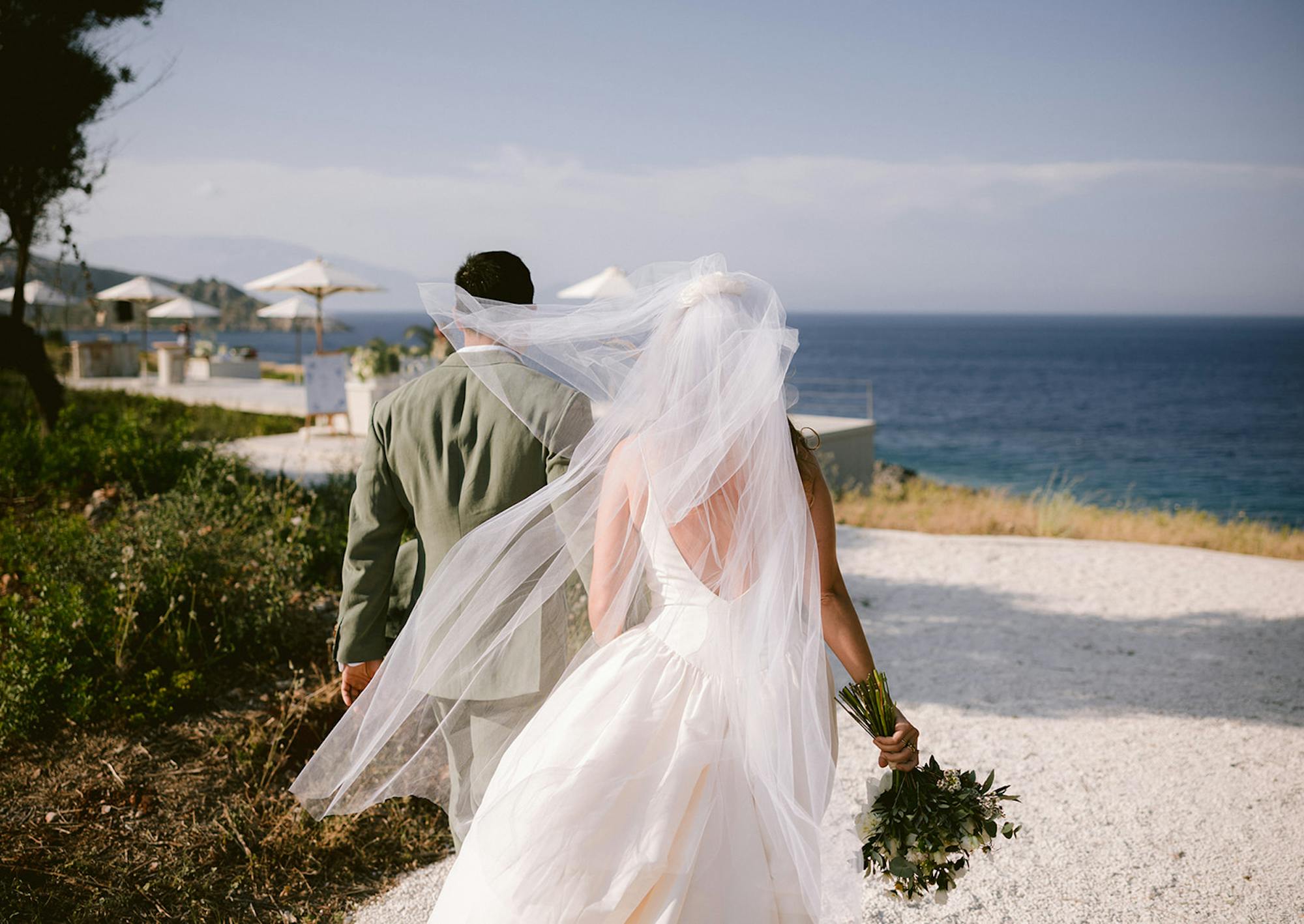 A newlywed couple photographed from behind as they walk away from their ceremony site, with the bride's voluminous white tulle veil caught dramatically in the wind, billowing across both figures. The groom wears an olive green suit while the bride in her white gown carries a lush green bouquet. They walk along a white stone path overlooking the deep blue Mediterranean Sea. In the distance, cream-coloured market umbrellas and wedding venue areas are visible on the clifftop, with misty blue hills and coastline stretching across the horizon. The romantic, windswept moment captures the joy of their seaside celebration.