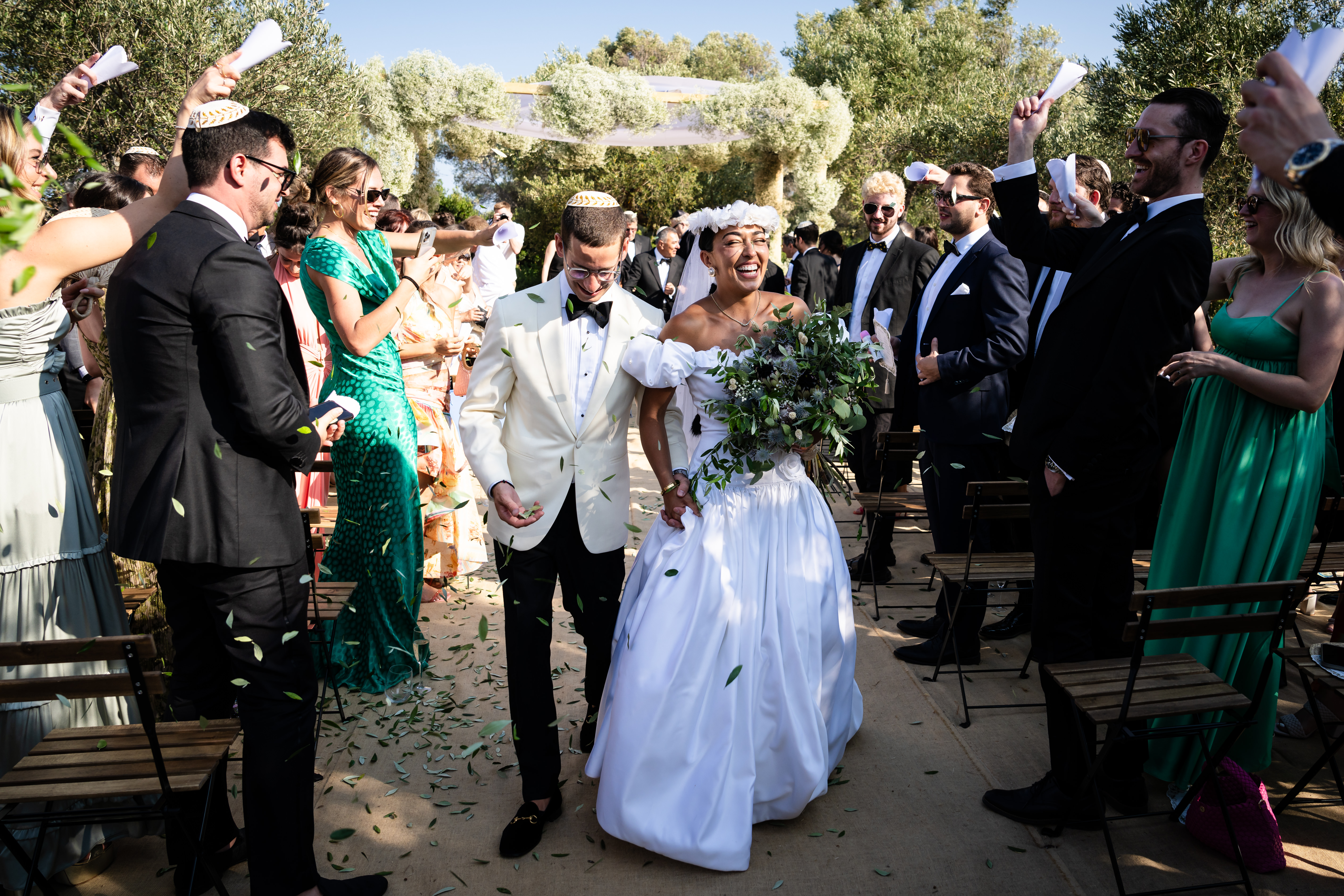 A jubilant recessional moment as the newlyweds walk down the aisle hand-in-hand through an outdoor ceremony space framed by lush olive trees. The groom wears an elegant ivory dinner jacket with black bow tie and trousers, while the beaming bride in a white off-shoulder ball gown with full skirt carries a cascading green foliage bouquet. Wedding guests line both sides of the aisle, waving white programs or napkins in celebration, creating a joyful tunnel of well-wishers. The guests are dressed in formal attire including black tuxedos and elegant gowns in emerald green and other colors. Behind them, a dramatic floral installation covered in white flowers and greenery creates an arbor over the ceremony seating area, with olive groves visible in the background under bright Mediterranean sunlight.