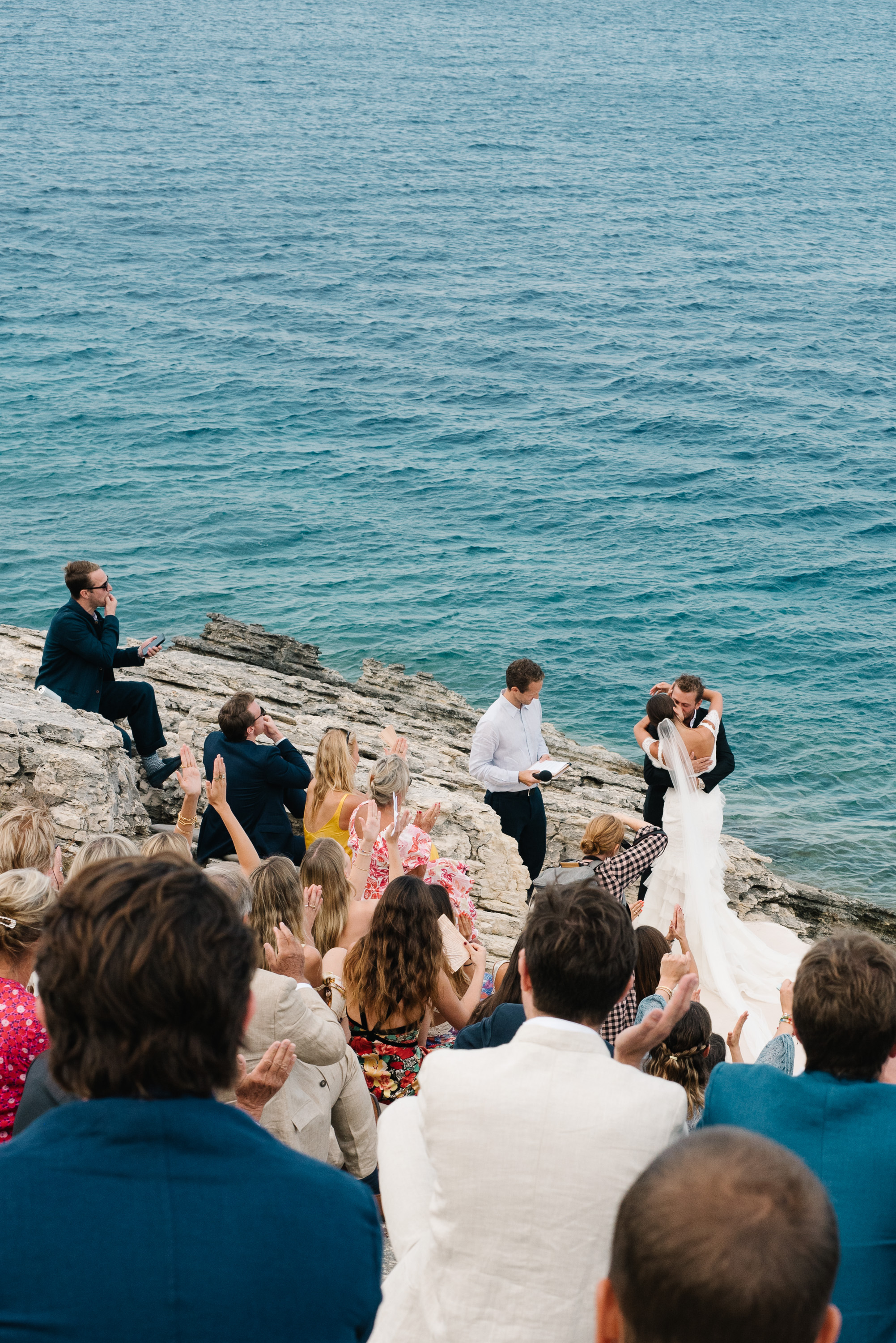 An intimate clifftop wedding ceremony photographed from behind the seated guests, looking down toward the couple and officiant standing on weathered limestone rocks at the water's edge. The bride in white and groom face each other as the officiant conducts the ceremony, with the vast turquoise-blue sea stretching to the horizon behind them. Guests seated on the rocky terrain wear a mix of formal and semi-formal attire, with one guest perched higher on the rocks to the left. The dramatic natural setting creates an unforgettable backdrop for this seaside celebration.