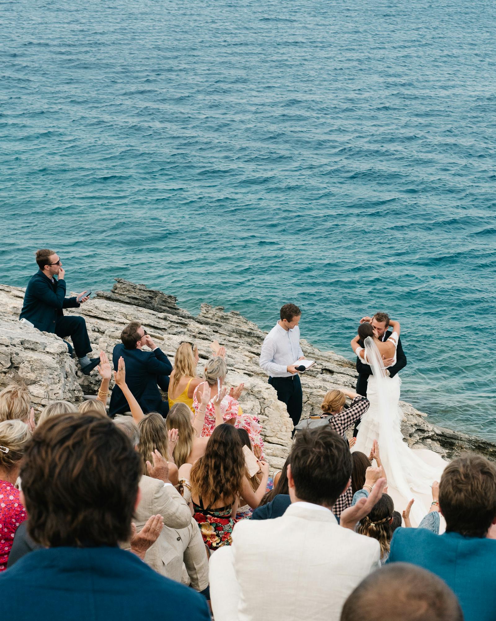 An intimate clifftop wedding ceremony photographed from behind the seated guests, looking down toward the couple and officiant standing on weathered limestone rocks at the water's edge. The bride in white and groom face each other as the officiant conducts the ceremony, with the vast turquoise-blue sea stretching to the horizon behind them. Guests seated on the rocky terrain wear a mix of formal and semi-formal attire, with one guest perched higher on the rocks to the left. The dramatic natural setting creates an unforgettable backdrop for this seaside celebration.