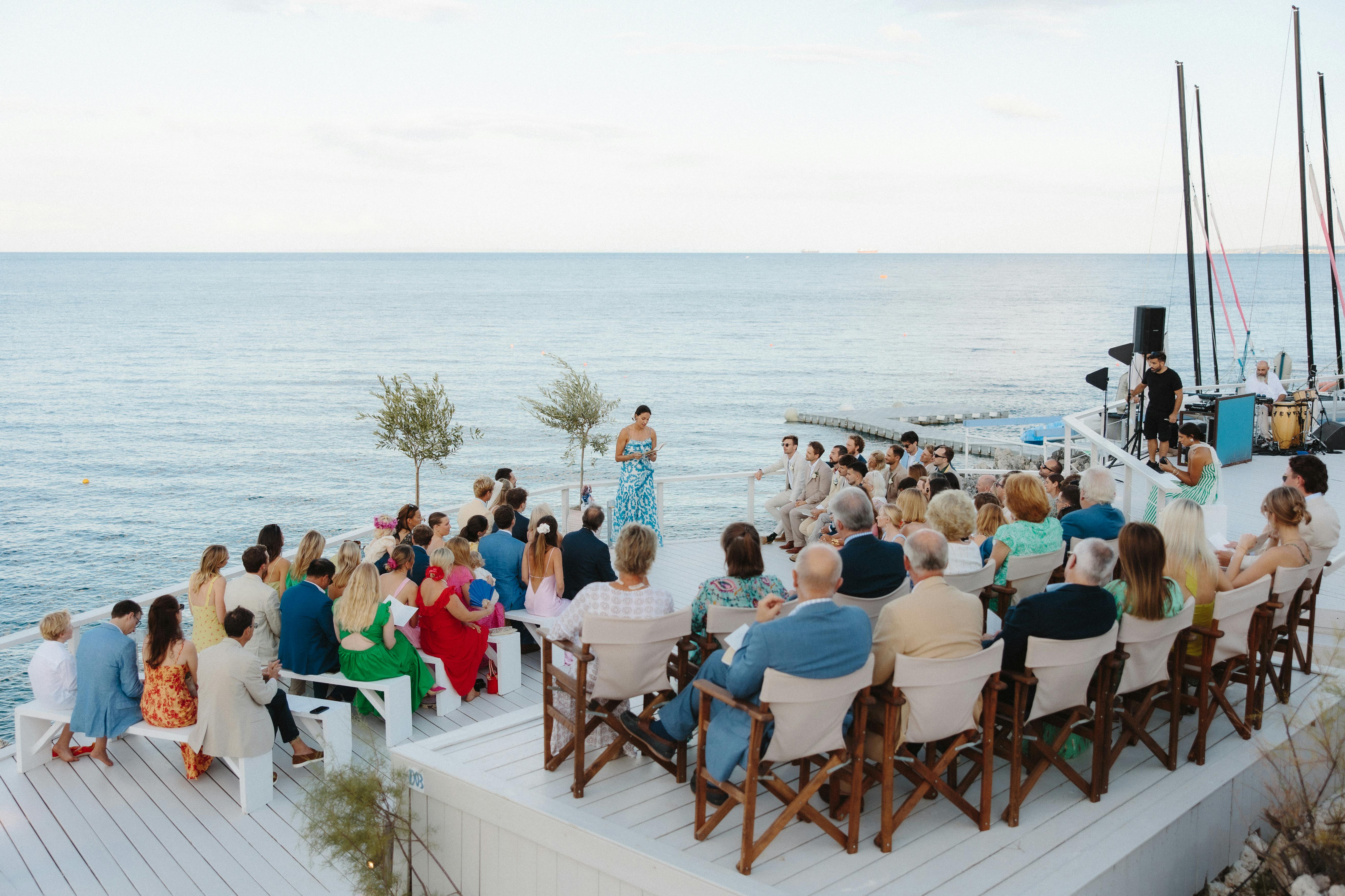 Wedding ceremony on a white wooden deck overlooking the sea. Guests in colorful summer attire sit on wooden chairs arranged in rows facing the ocean. An officiant in a blue floral dress stands before the couple and guests on the waterfront platform, with calm blue water and horizon visible in the background. Decorative tall wooden poles frame the ceremony space.