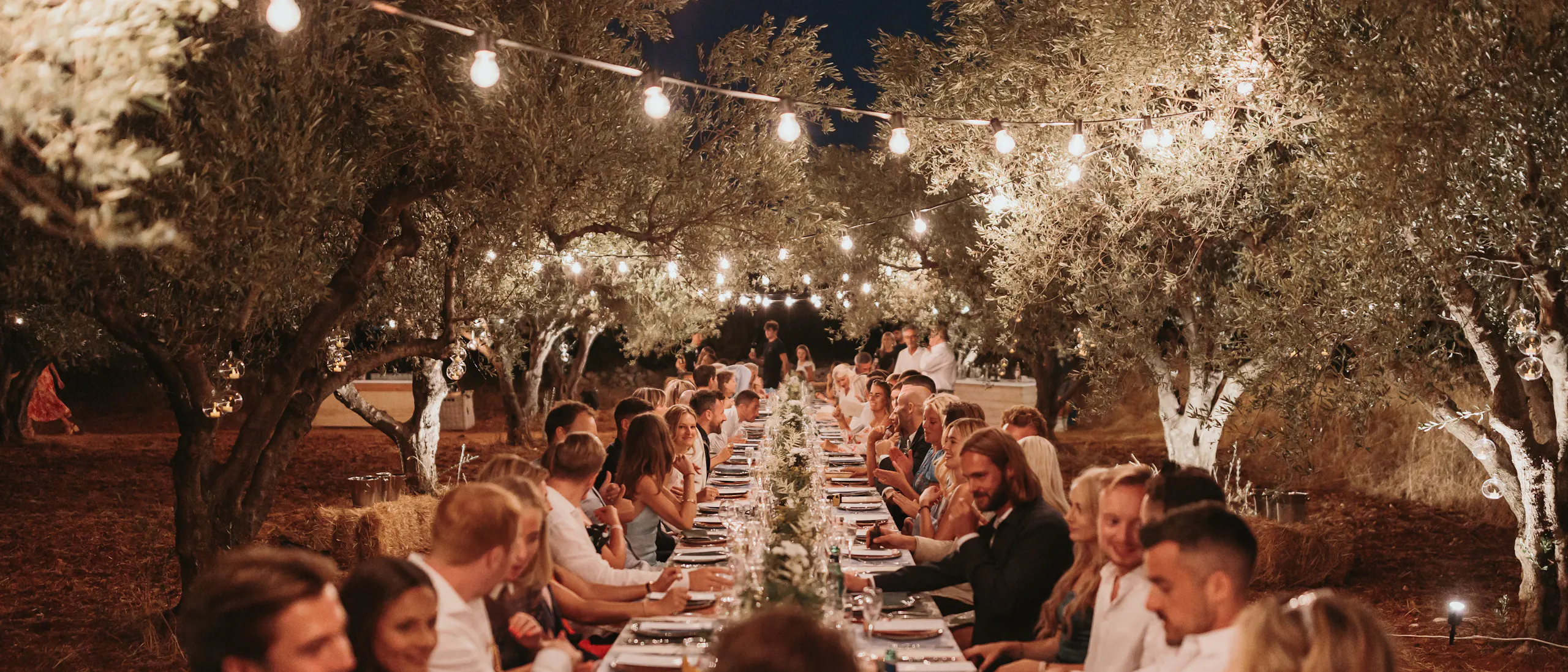 Evening wedding reception dinner in an olive grove with guests seated at a long table under string lights. The romantic outdoor setting features illuminated olive trees creating ambient lighting, with guests in formal attire enjoying dinner along both sides of the white-clothed table decorated with greenery runners.