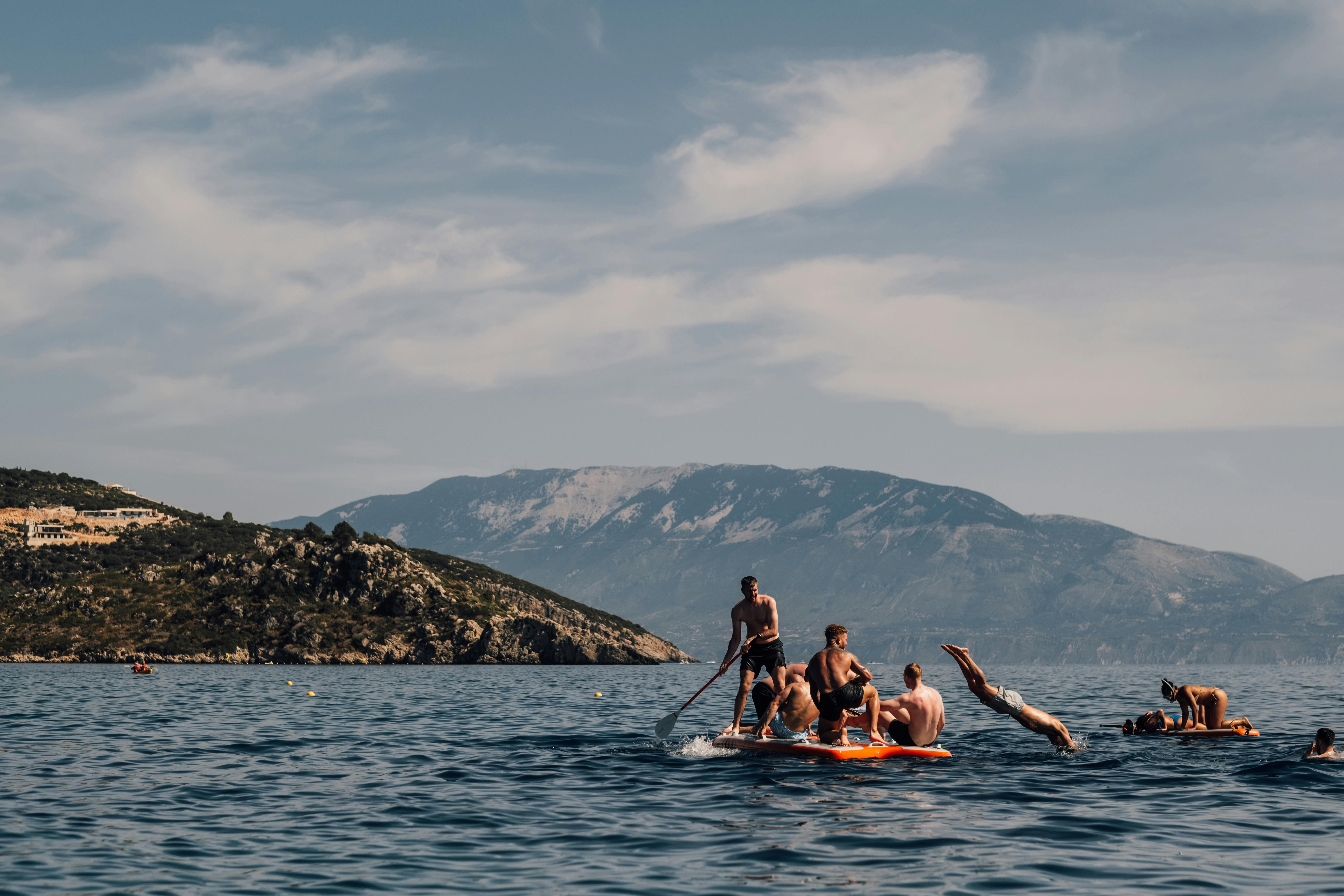 Group of people enjoying water activities on a large paddleboard in calm blue waters, with some diving off while others relax on board. Mountain ranges form a scenic backdrop across the bay, with rocky coastal hillsides visible on the left under a cloudy sky.