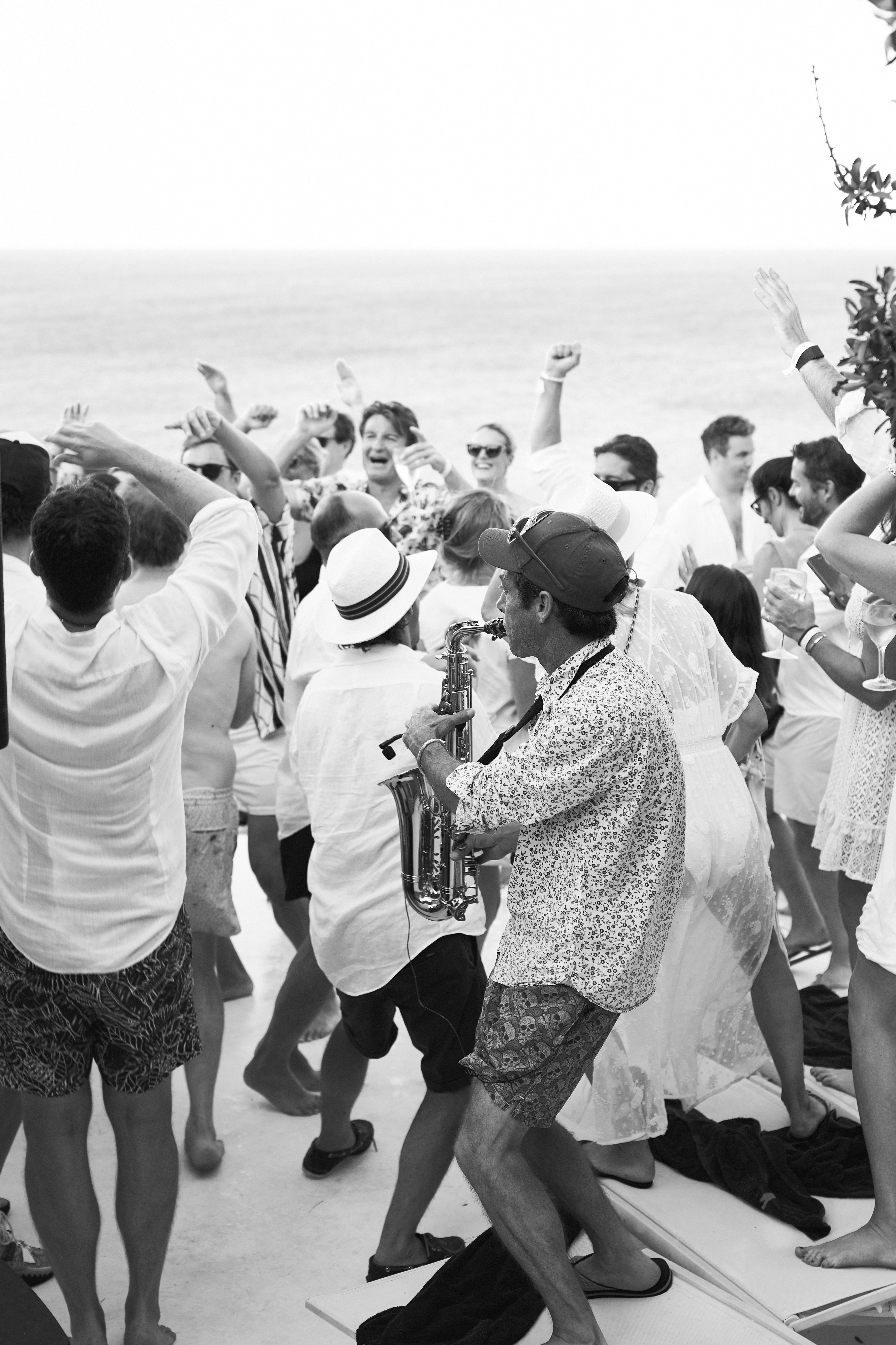 Black and white photo of wedding guests dancing outdoors with live saxophone musician. Guests in summer attire with raised arms dance energetically around a saxophonist in patterned shirt and straw hat, with the ocean visible in the bright, hazy background.